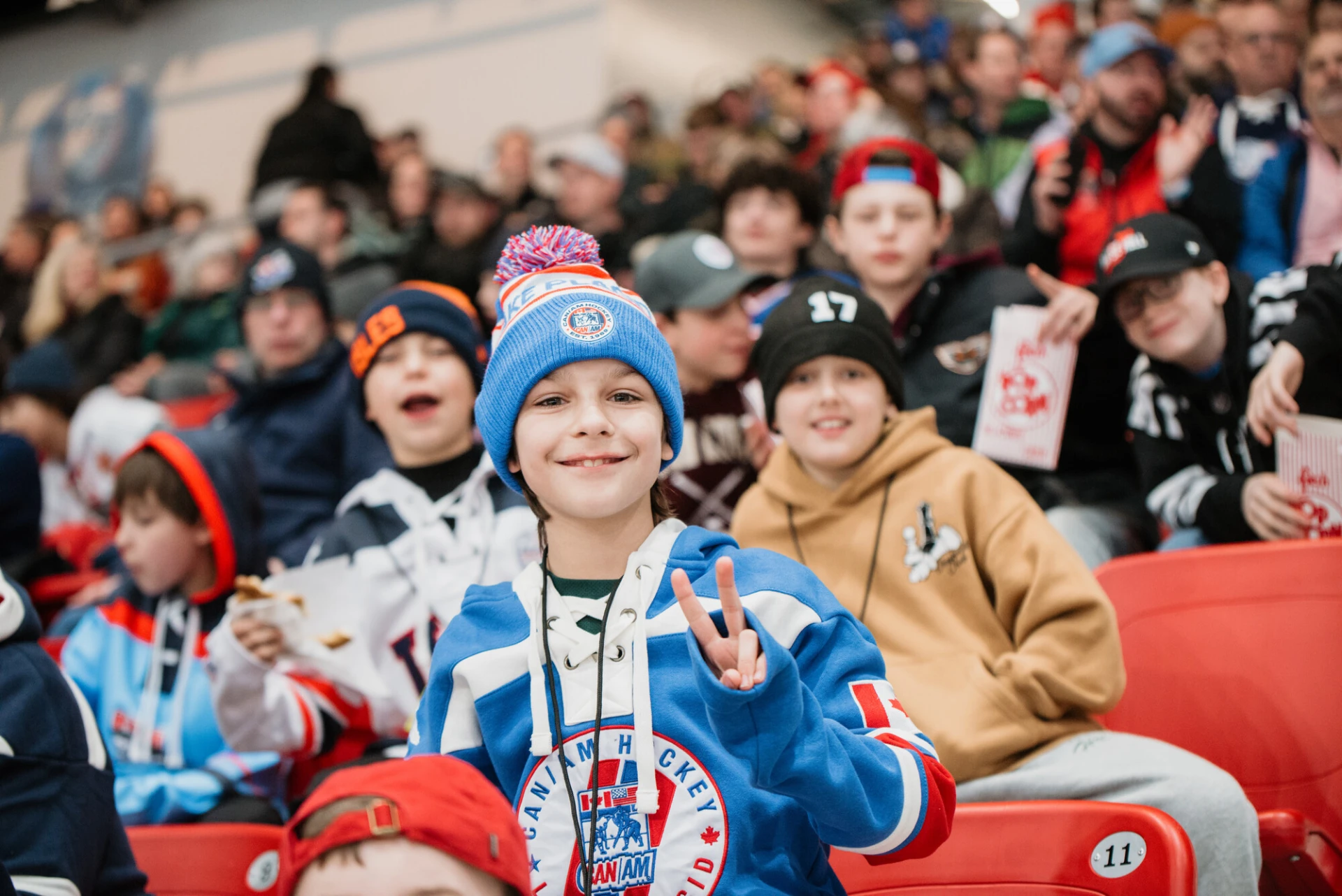 Child in hockey jersey holds up hand in peace sign while in stands of the 1980 Herb Brooks Arena along with a crowd of people