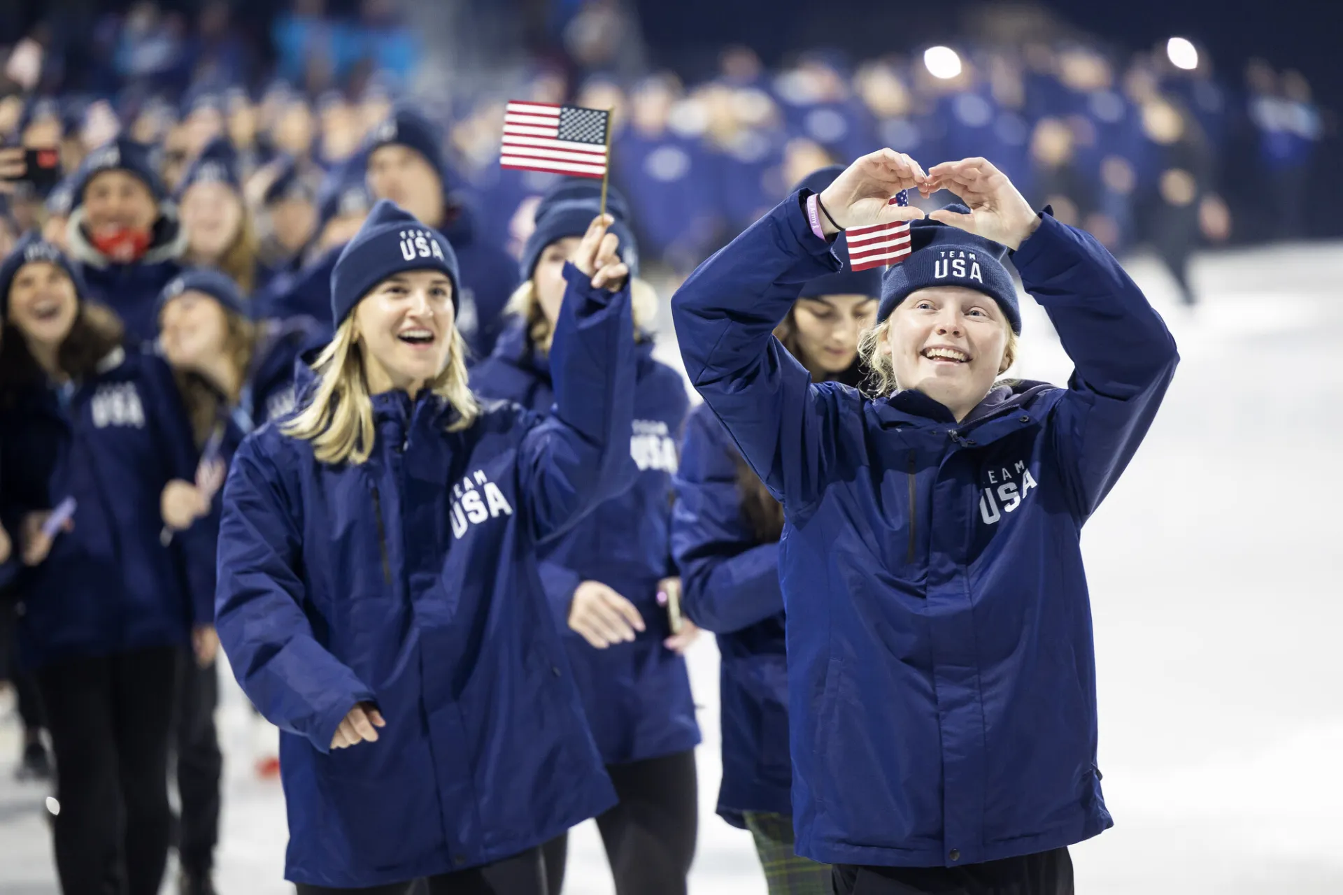 Team USA athletes wearing blue track suit uniforms and beanies walk out on ice during FISU opening ceremony inLake Placid. One girl waves a small American flag while another holds her hands up in the shape of a heart.