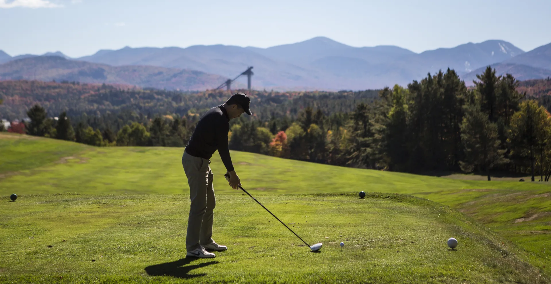 A golfer tees off with the Adirondack High Peaks and Olympic ski jumps in the background.