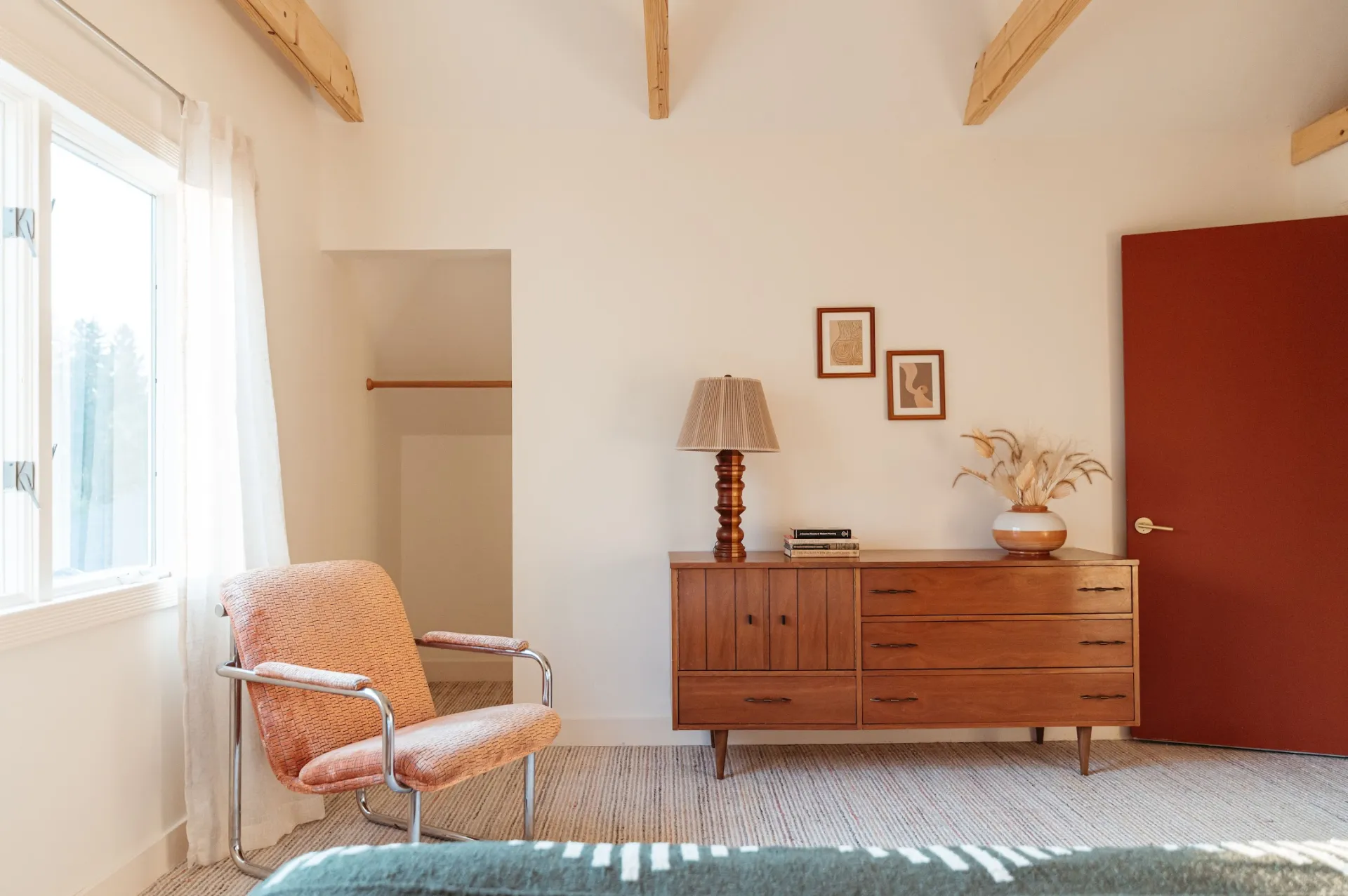 A retro pink upholstered and metal framed chair, large wooden dresser with unique lamp and fluffy grasses in a vase.