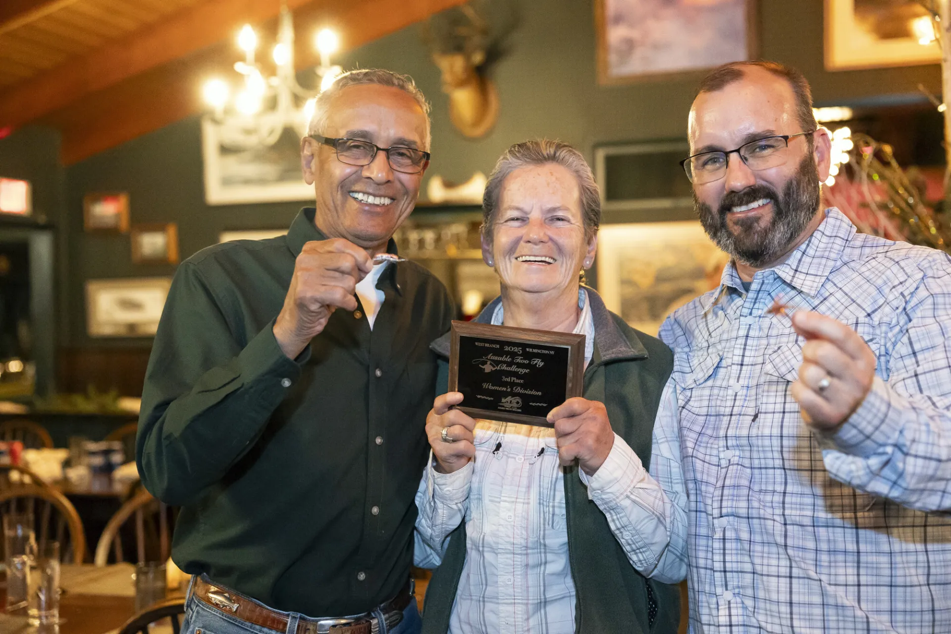 Two men holding up fishing flies stand on either side of a woman holding a plaque at an Ausable River Two-Fly Challenge event