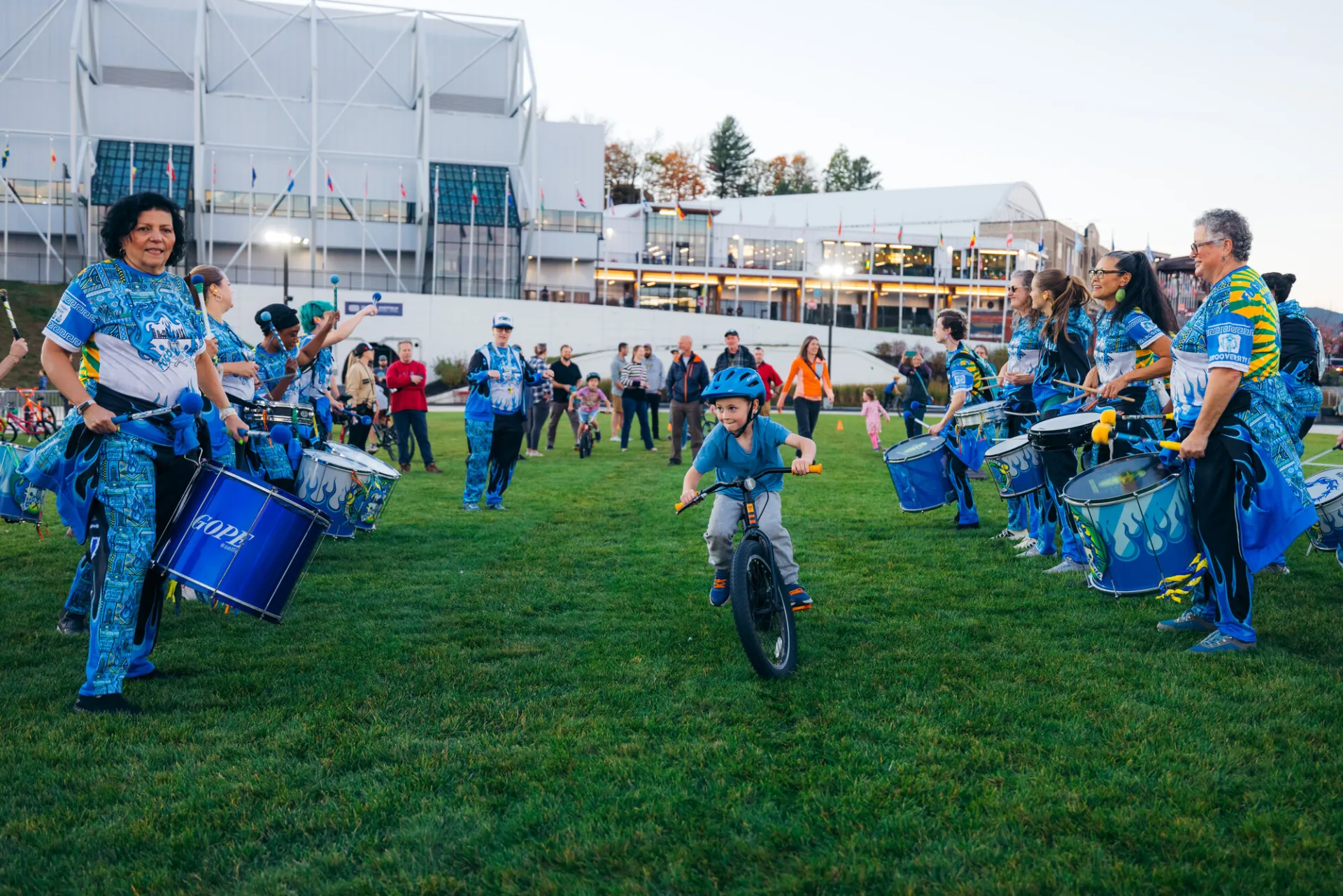 Child rides bike through aisle created by drum line on grass of Olympic Speed Skating Oval