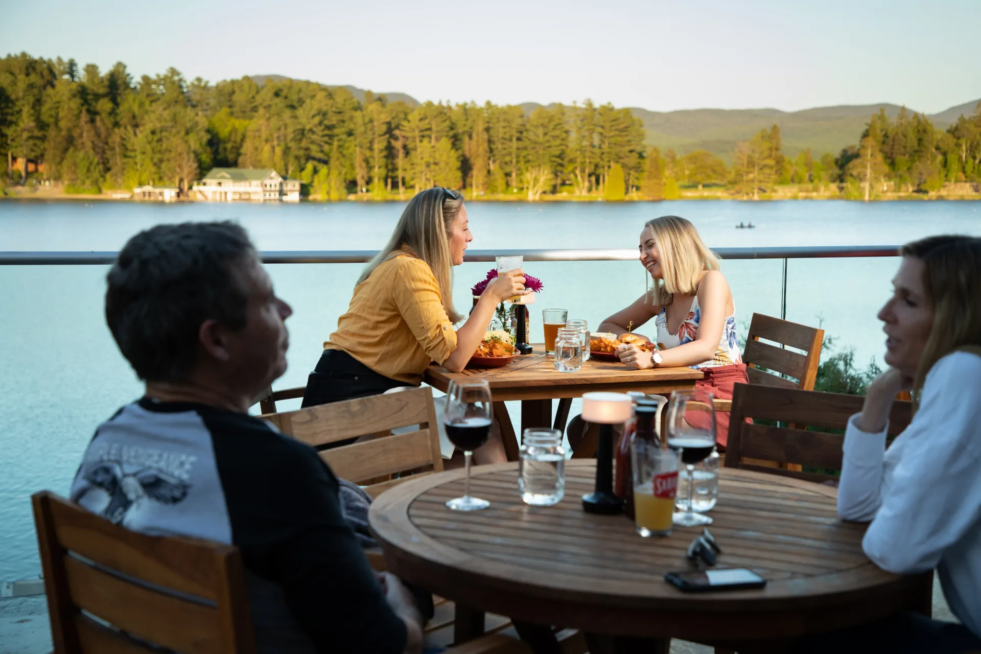 Two women site across from each other at bistro table on deck overlooking lake drinking cocktails with other diners in the foreground
