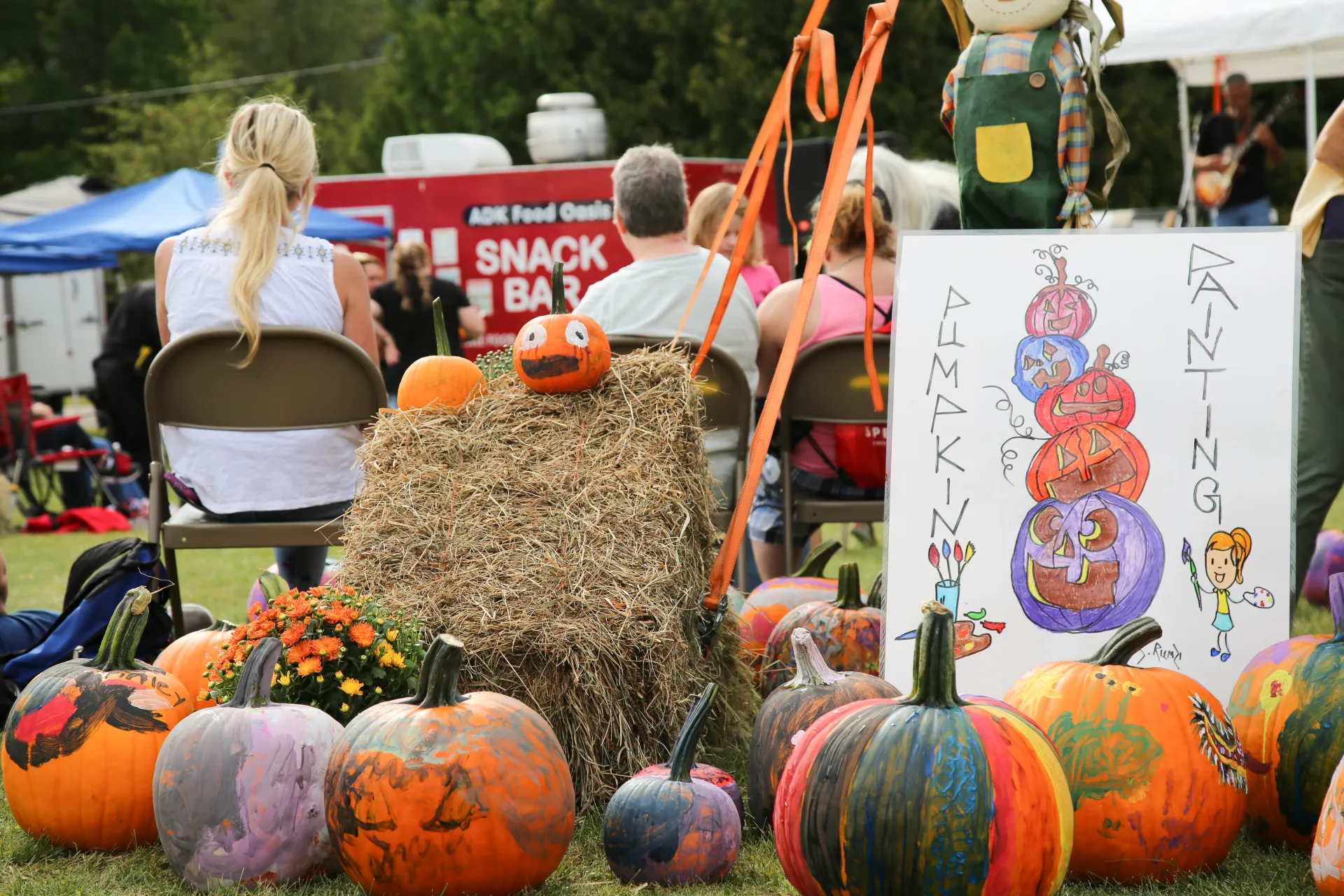 Collection of painted pumpkins and hay bales next to a tent pole and paint a pumpkin sign
