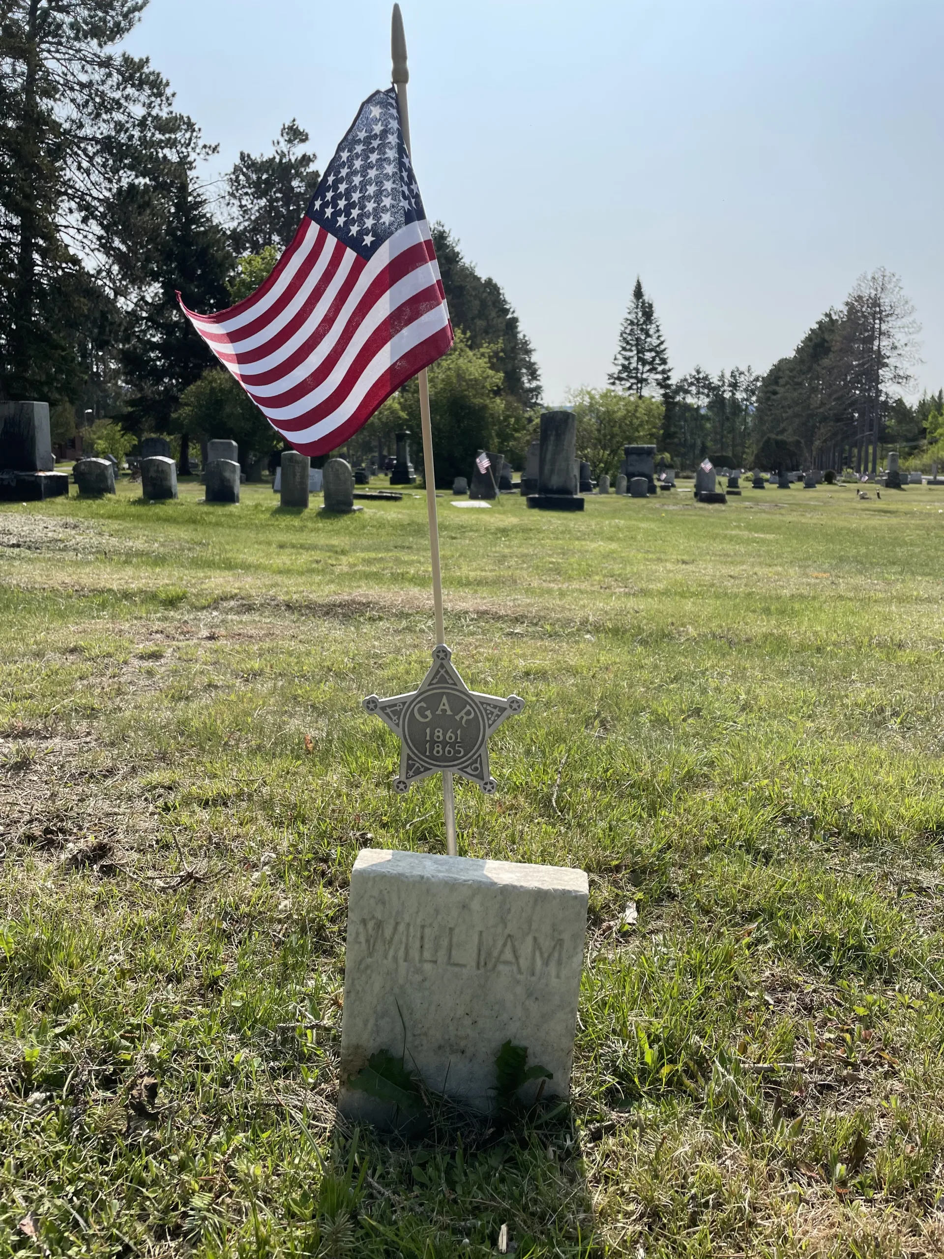 A small gravestone amid green grass with the name William and a metal star stick into the ground behind it that says GAR 1861 and 1865 and above that a small American flag