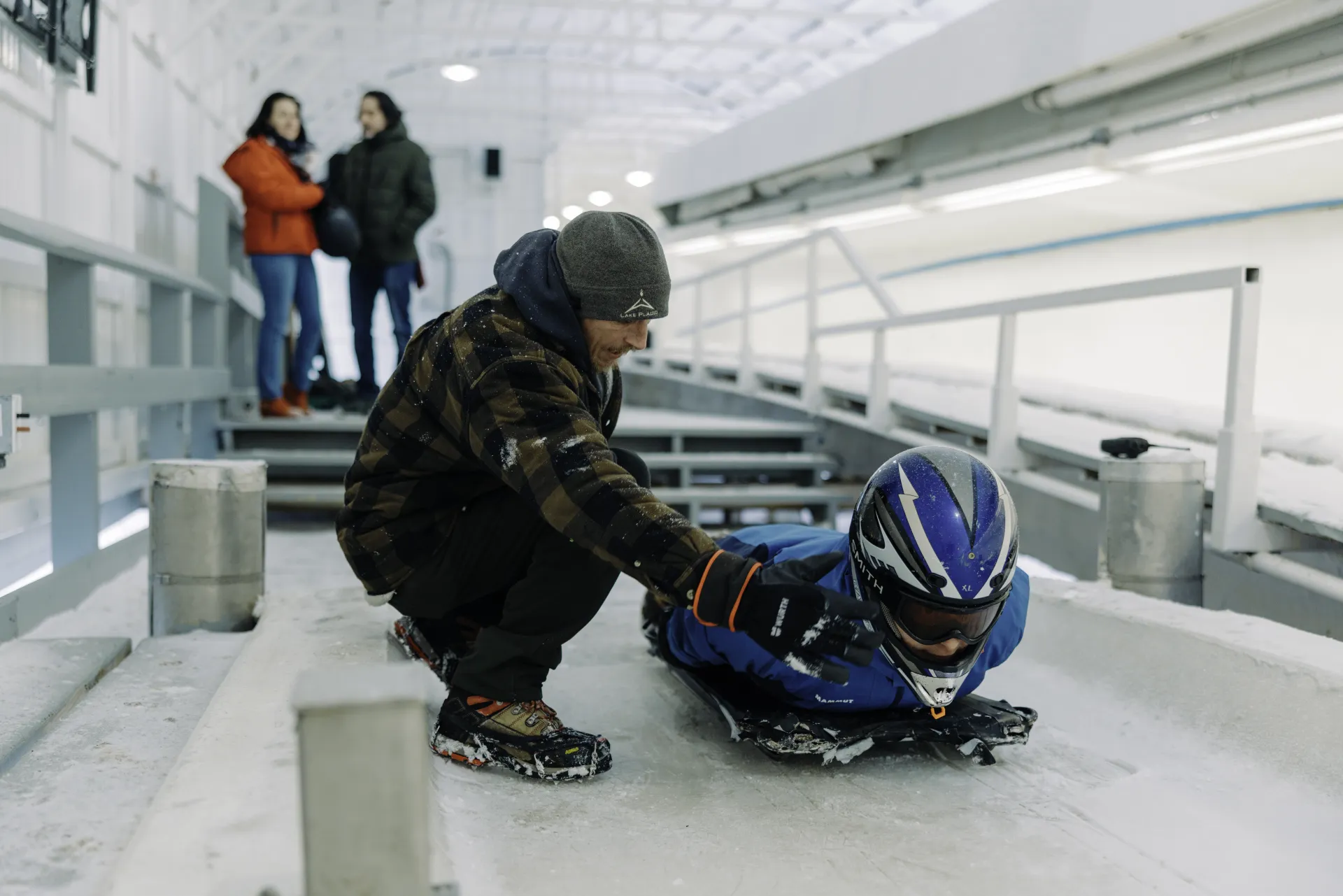 Track worker provides instruction to person in helmet laying on skeleton sled on bobsled, luge, and skeleton track