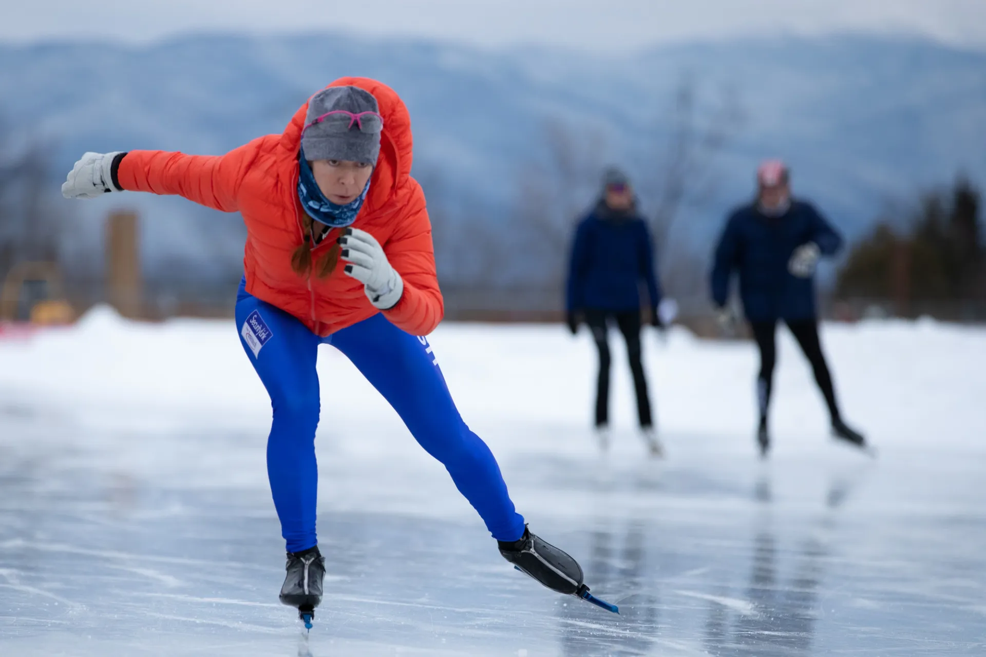 Person in red puffy coat and bright blue spandex pants wearing a hat and white cloves speed skates and a crouched position on an outdoor skating oval. 