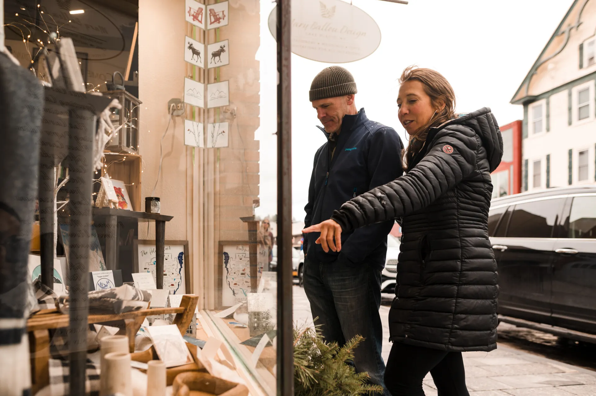 Man and woman in winter clothing look into a shop window with holiday decor and stationary. The woman is pointing at something and the perspective is from the inside of the window.