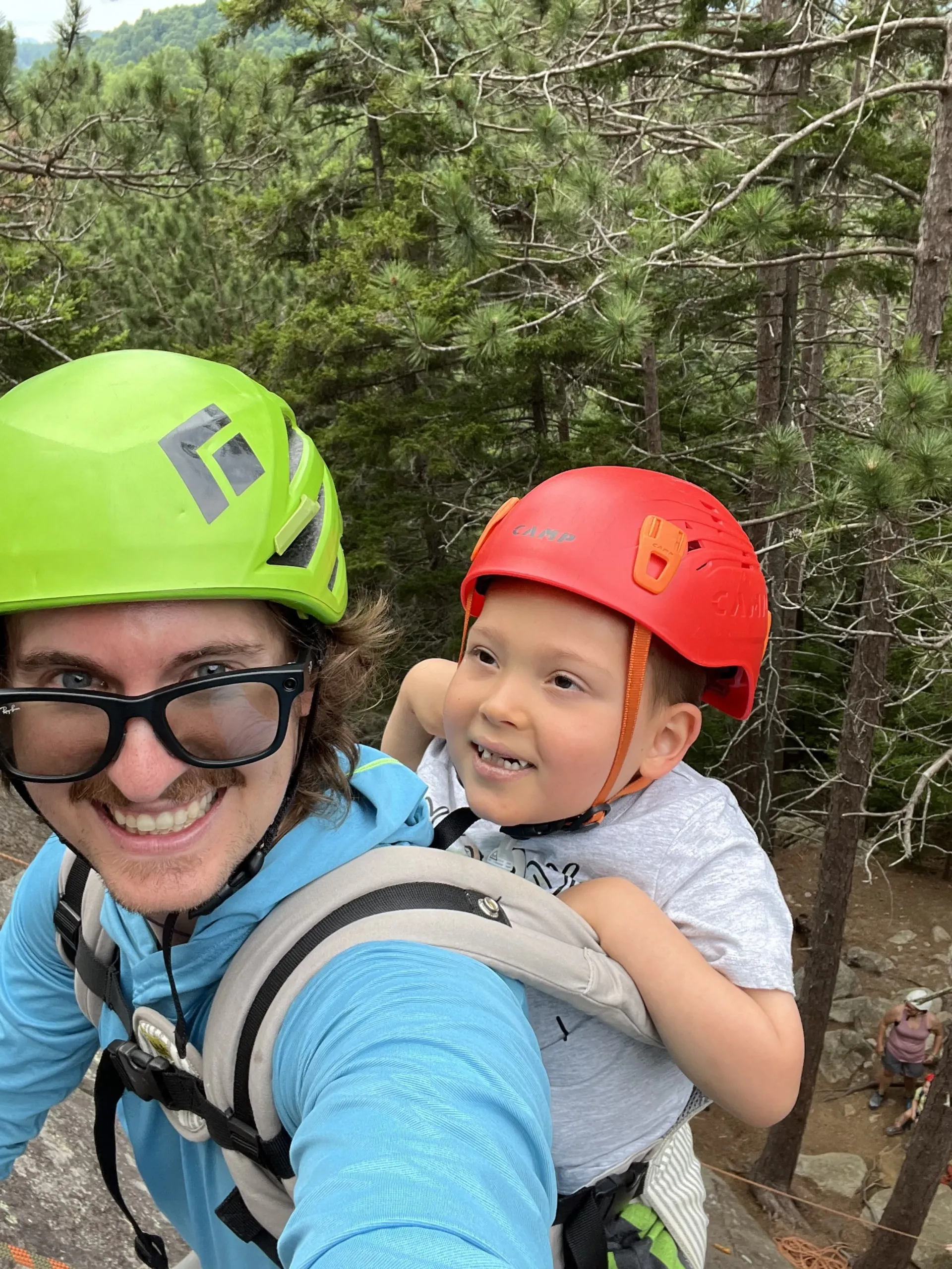 A smiling man faces camera while carrying a young child in a back pack, both donning helmets.