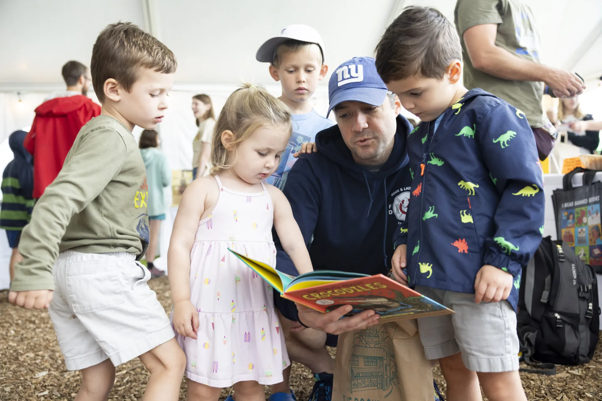 Three small children gather around a man crouching and reading a book