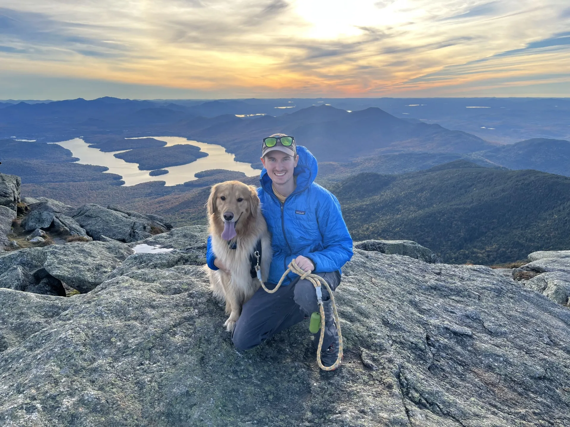 A man and his golden retriever sit atop a summit overlooking Lake Placid at sunset