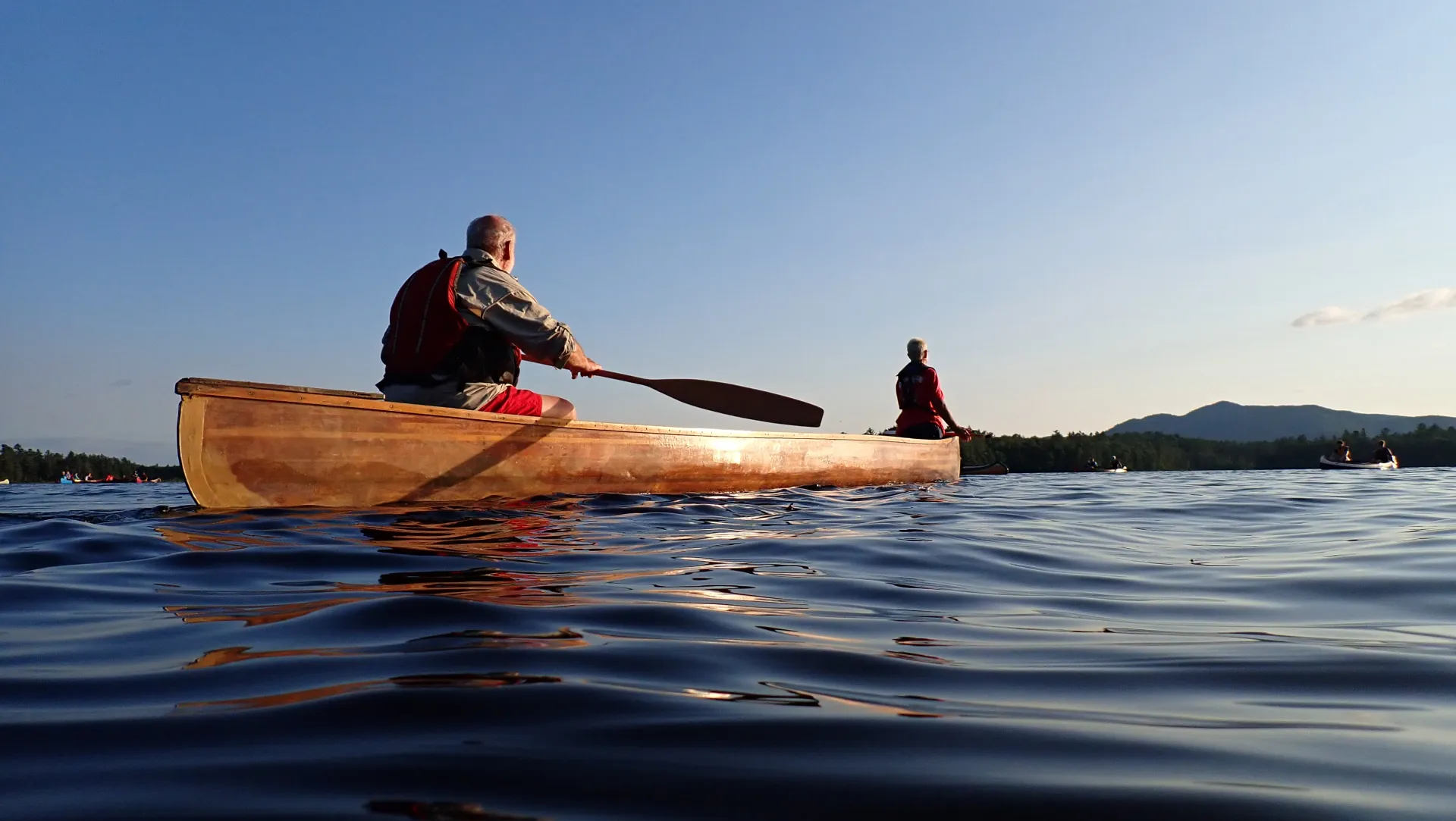 Sun reflects off the high gloss finish of a wooden canoe paddled by an older couple.