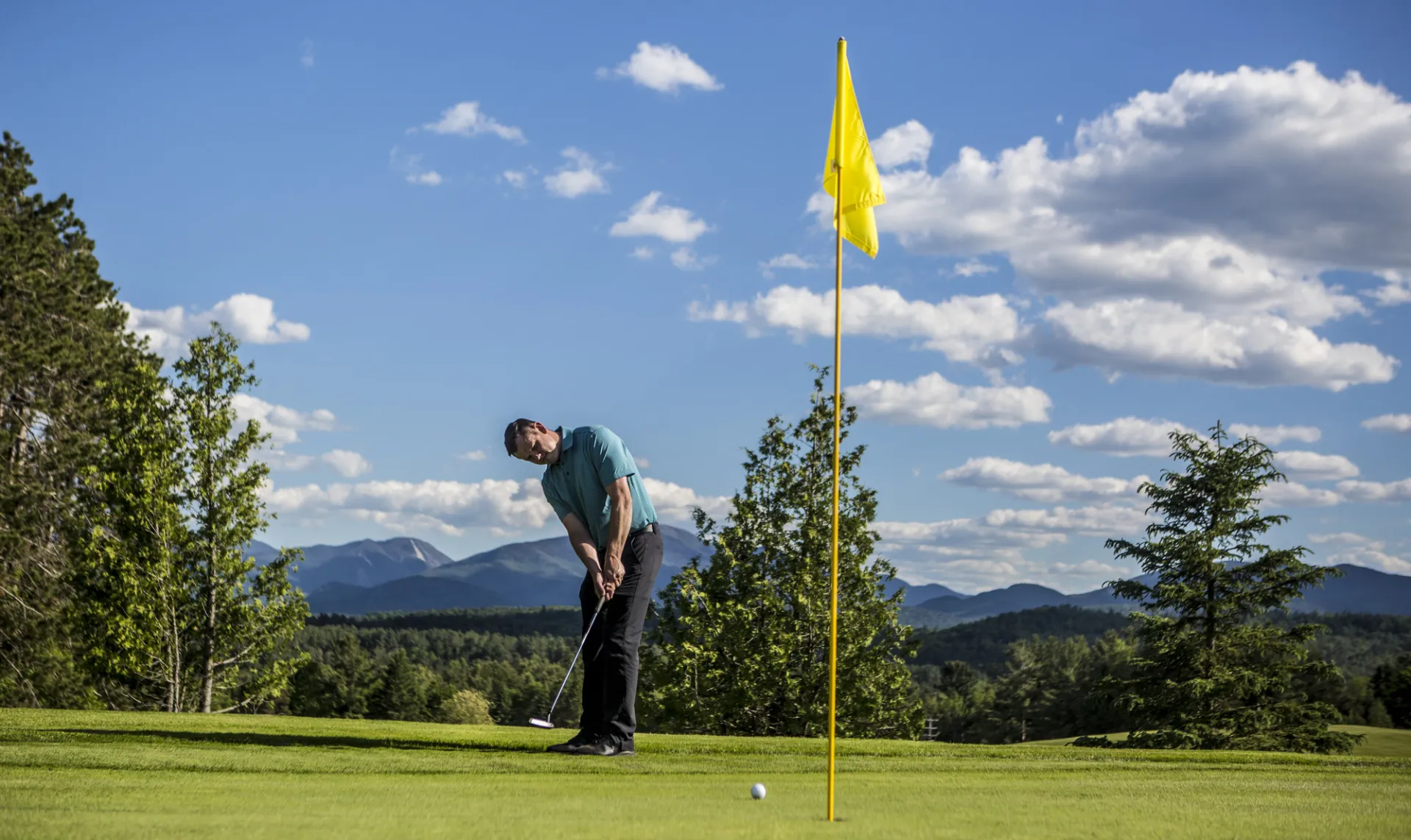 A man putts a golf ball toward the pin on a course with large mountain views in the background.