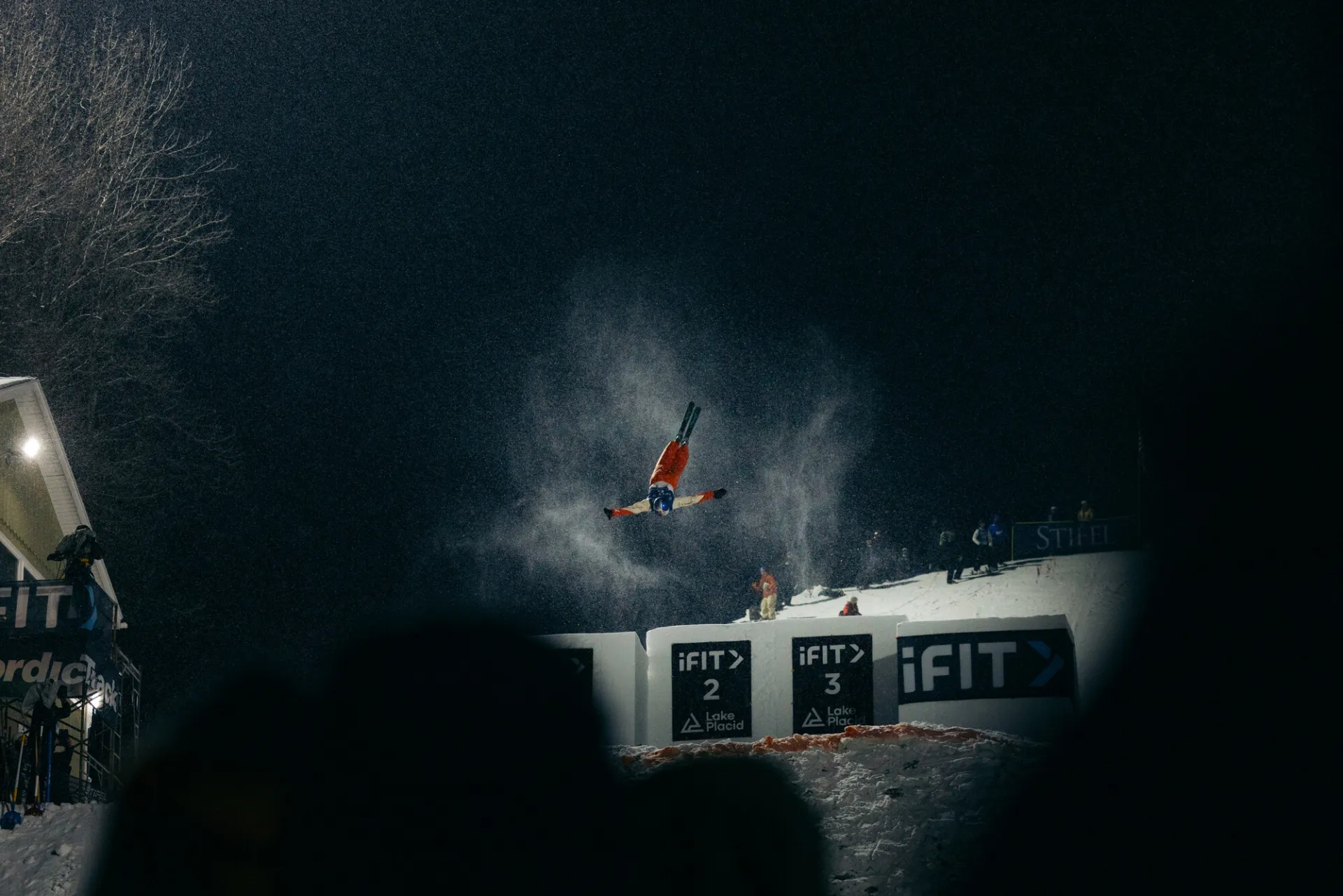 Freestyle aerialist skier performs a flip after skiing off a jump at a nighttime competition under bright lights while a crowd looks on