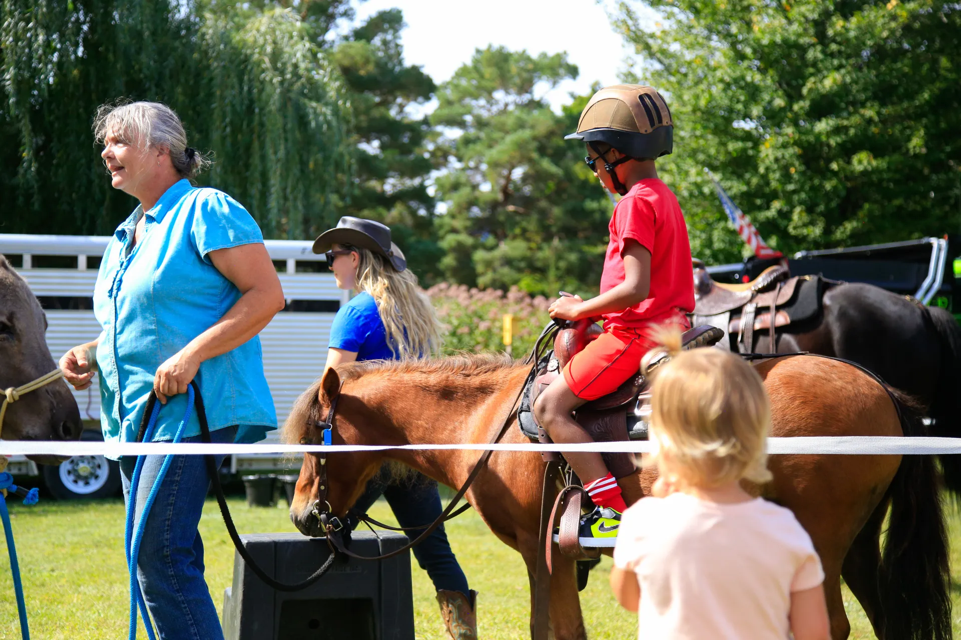 Young boy dressed all in red wearing a black helmet rides a small horse led by an older woman in a blue shirt.