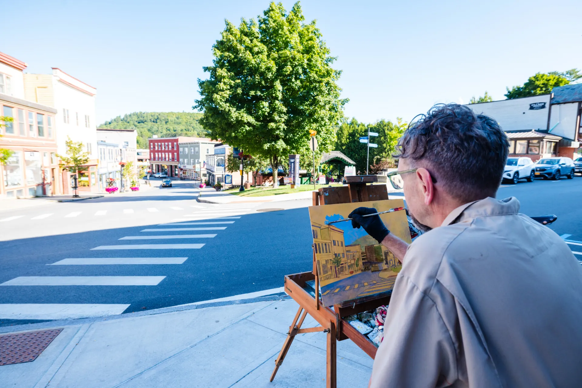 An artist stands at an easel on a sunny sidewalk, painting a plein air landscape of Saranac Lake.