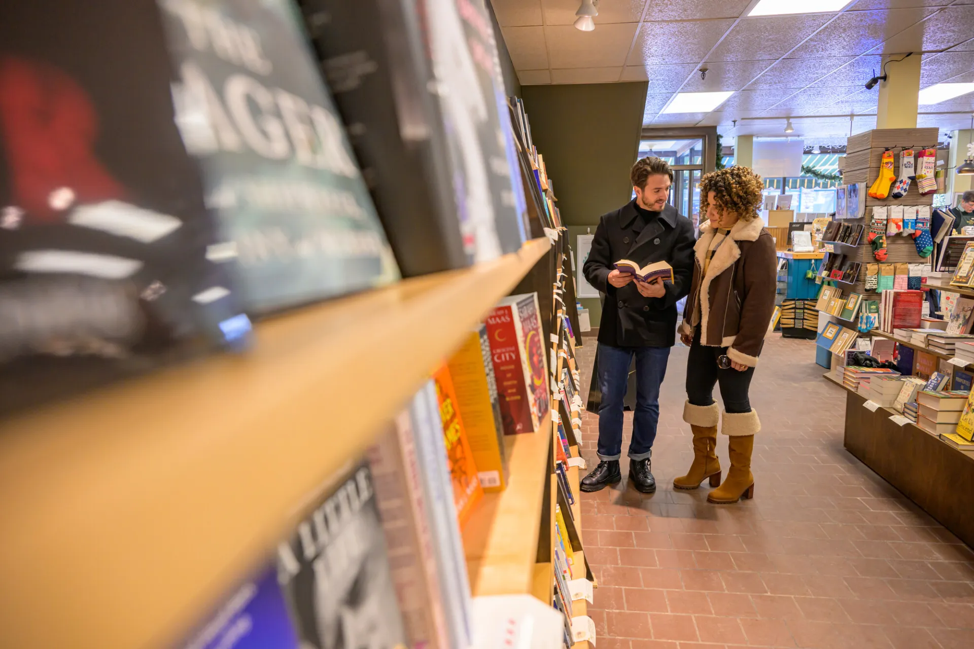 Couple in winter outerwear stands at bookshelves in bookstore looking at paperback