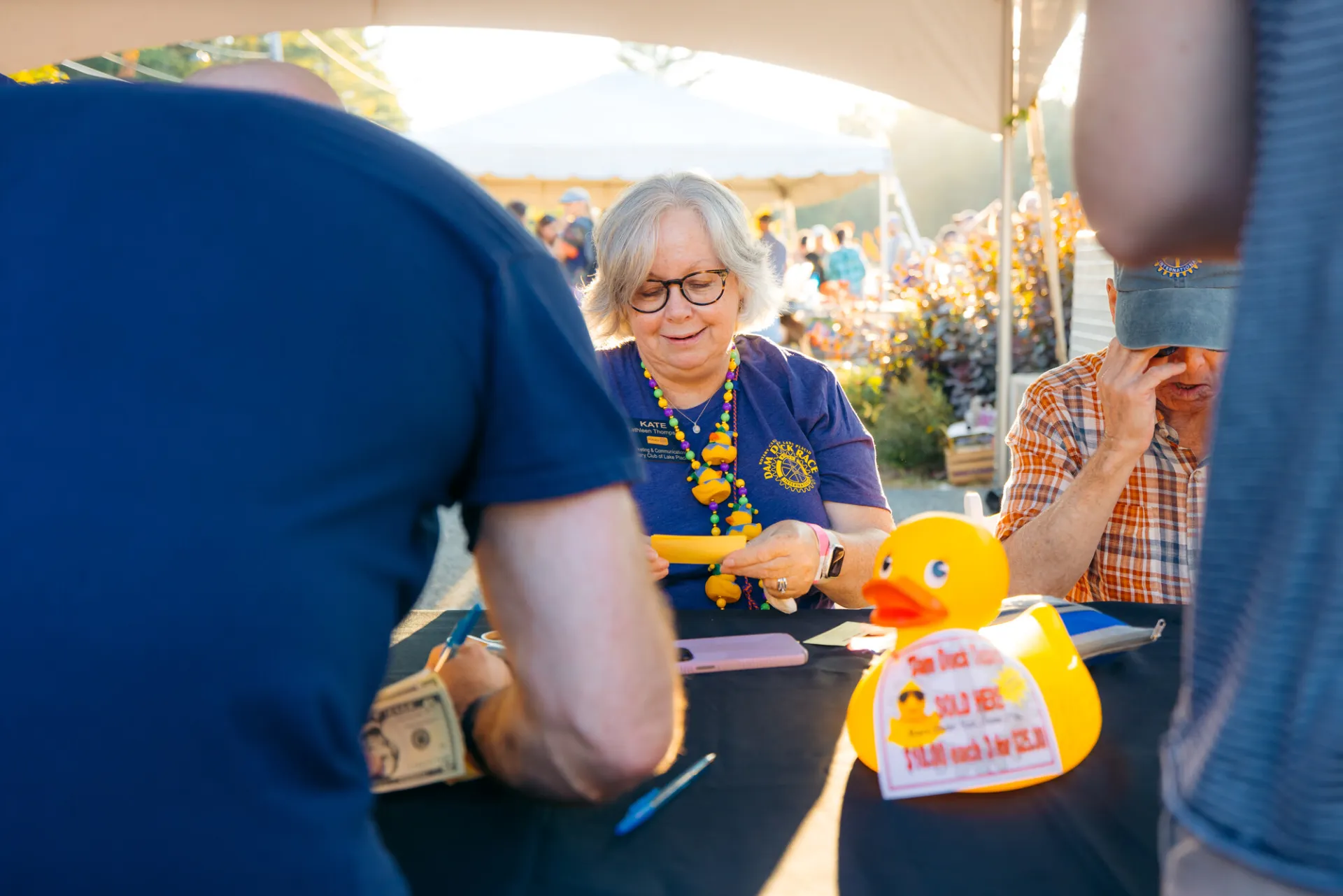Woman with grey hair and glasses wearing blue Rotary shirt and duck accessories is seen selling something 