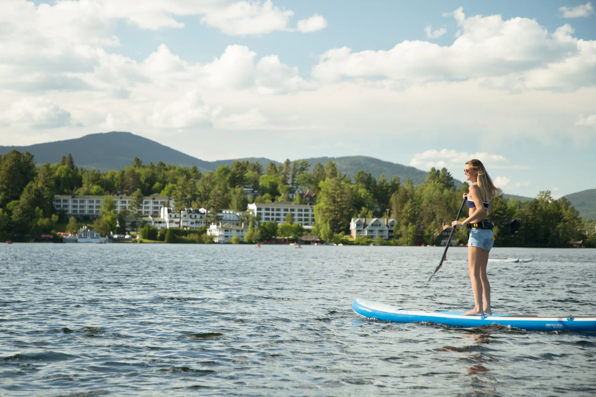 A woman paddles a stand up paddleboard on a lake with a town and mountains in the background.