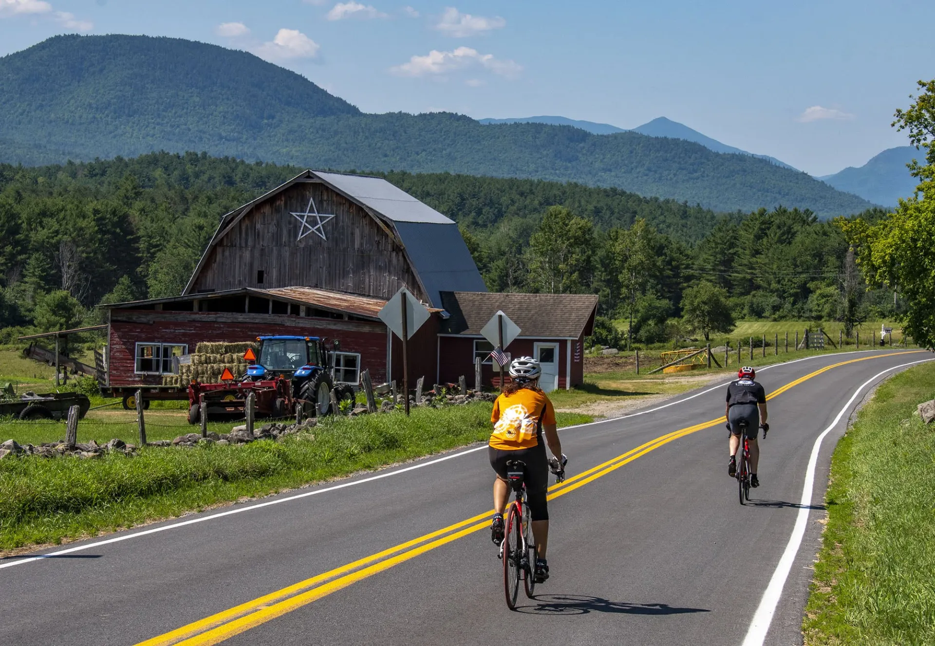 Two cyclists ride on a rural road past a barn and tractor, surrounded by lush green hills and mountains, under a clear blue sky.