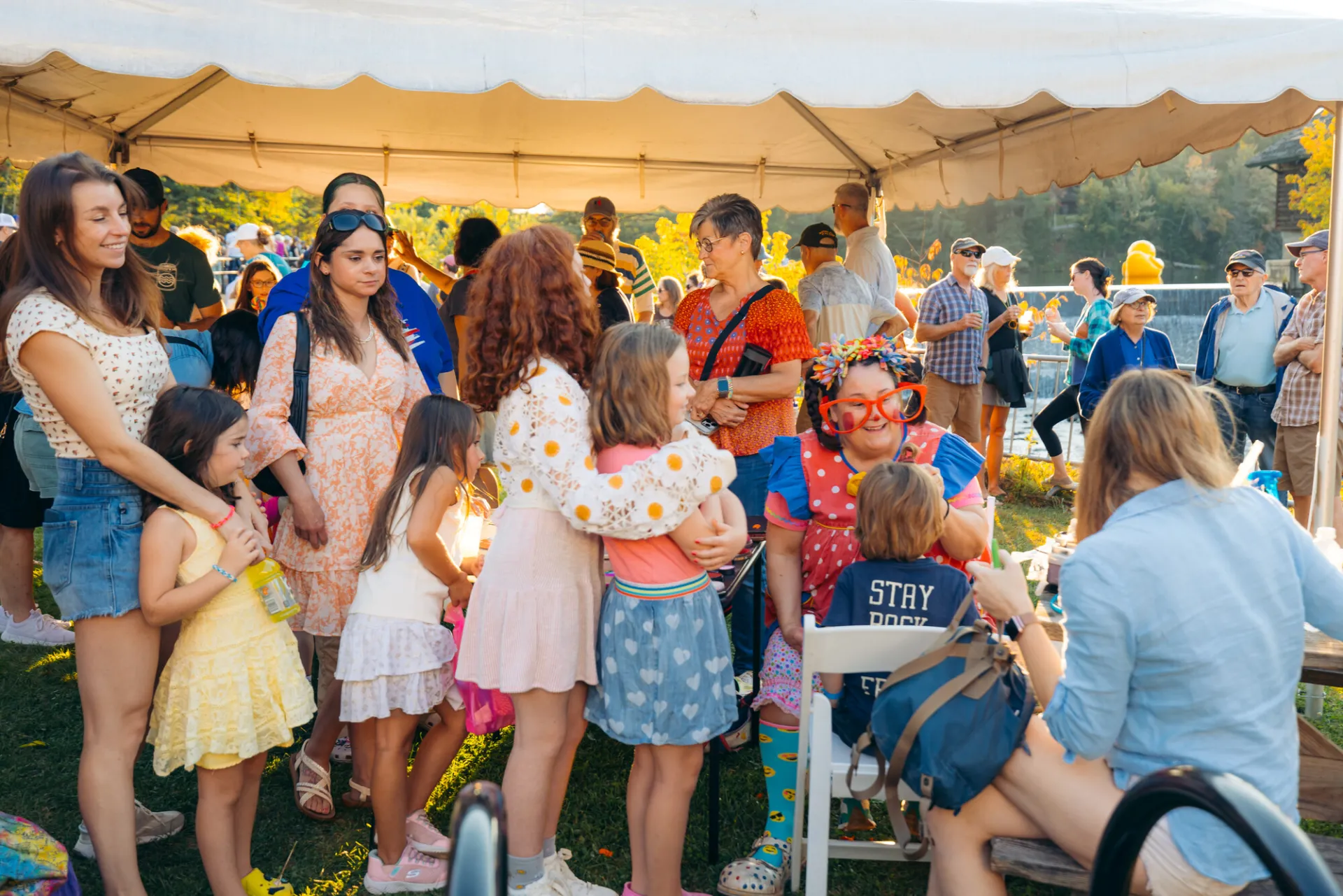Children and parents wait in line looking on as seated clown paints the face of a child 
