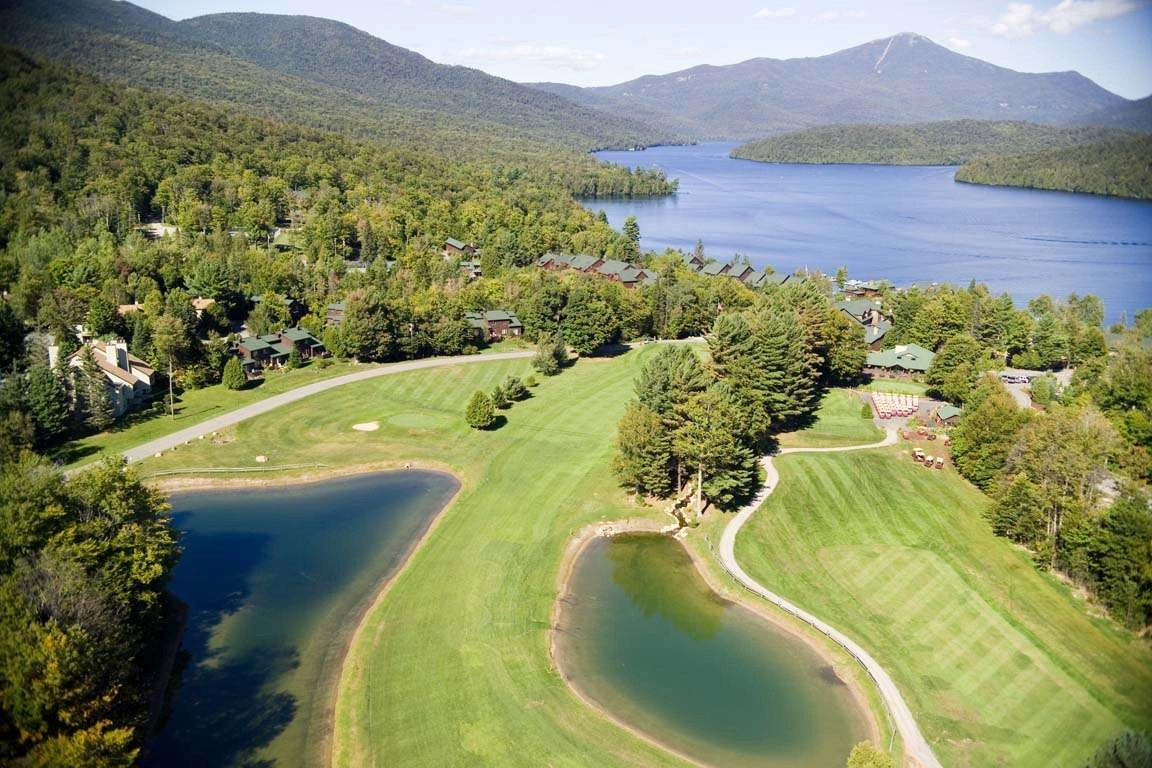 An aerial view of the pristine grass of the course by the deep blue Mirror Lake and mountains of the High Peaks surrounding.