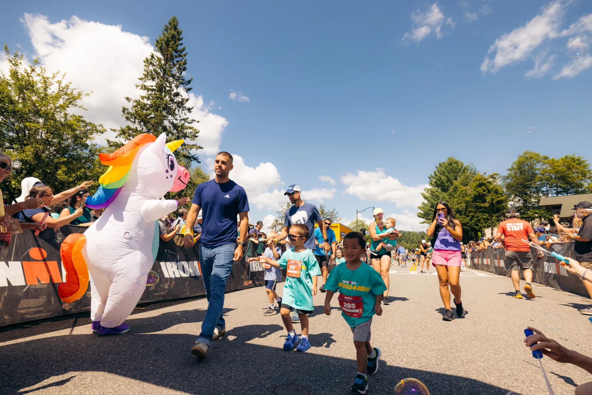 Group of kids and parents, some filming, walk in Ironkids race course past inflatable unicorn mascot