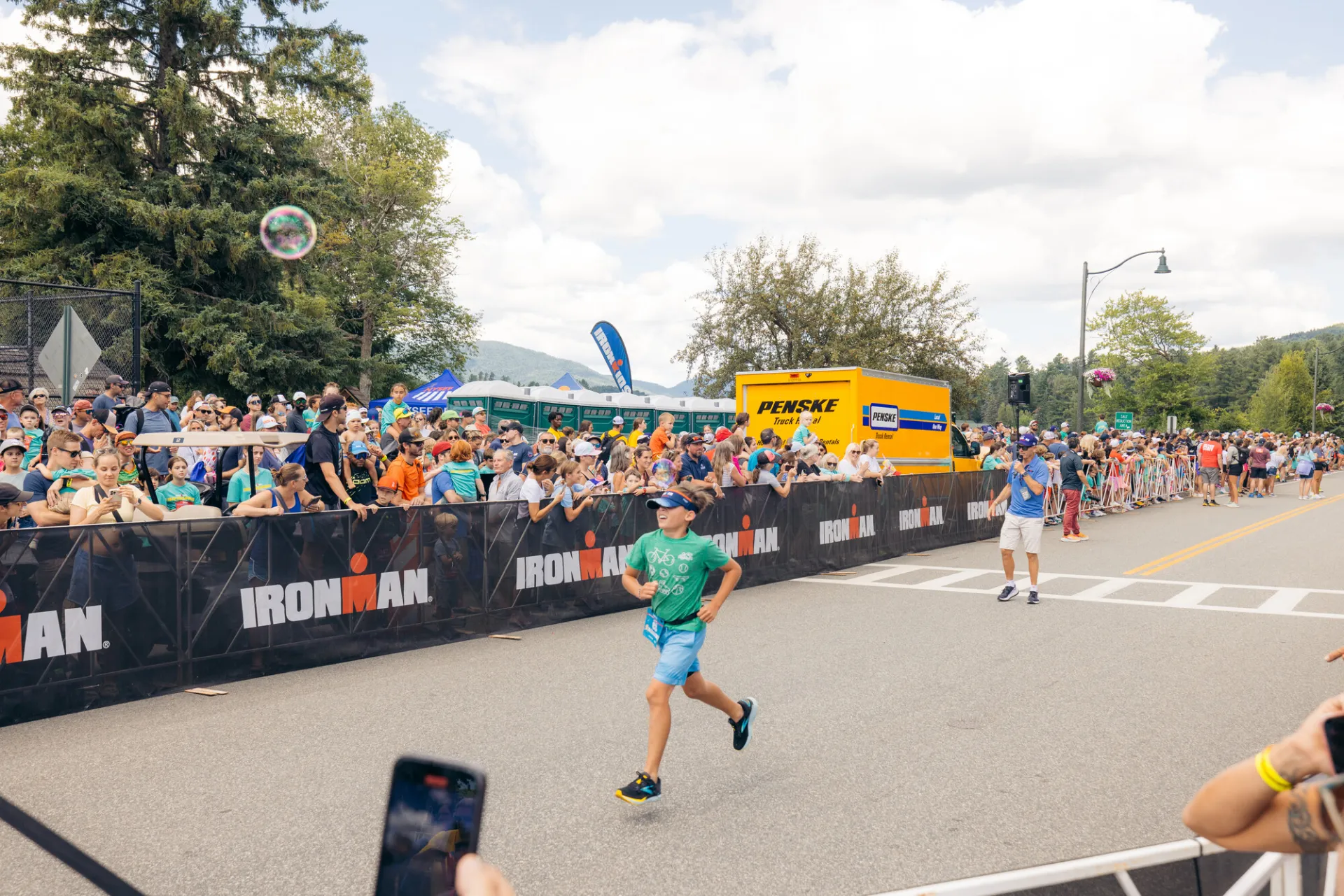 Child in teal shirt, blue shorts, and visor runs on Ironkids course while spectators cheer from behind Ironman fencing