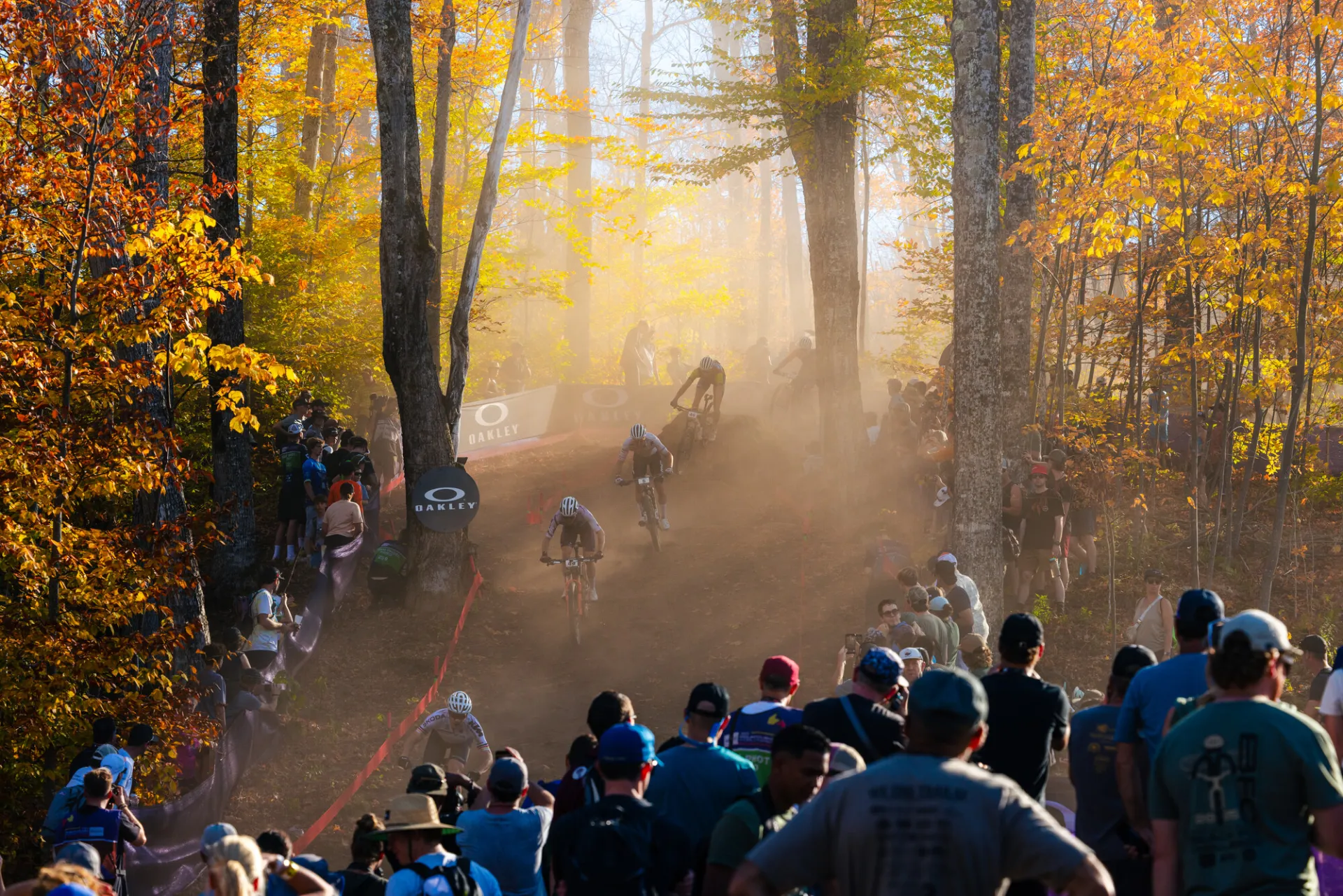 Dust flies and catches light through trees as mountain bikers race down cross-country course and spectators look on