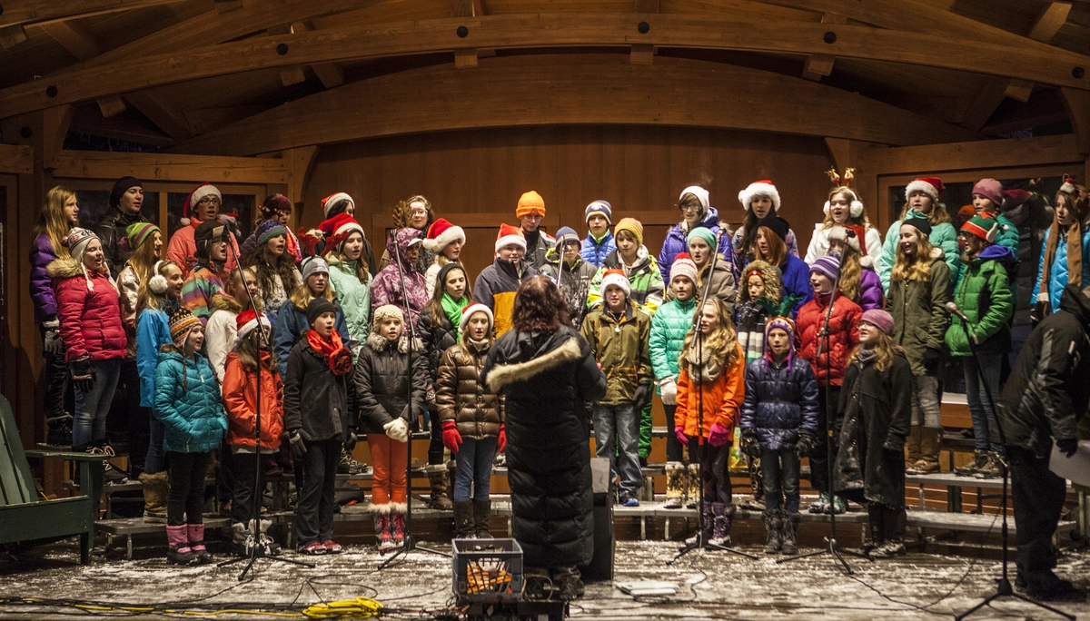 Several rows of children in winter and Santa hats sing in a lit, outdoor bandshell.
