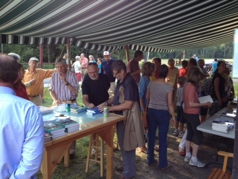 A group of people gather under a striped awning, browsing books displayed on a table.