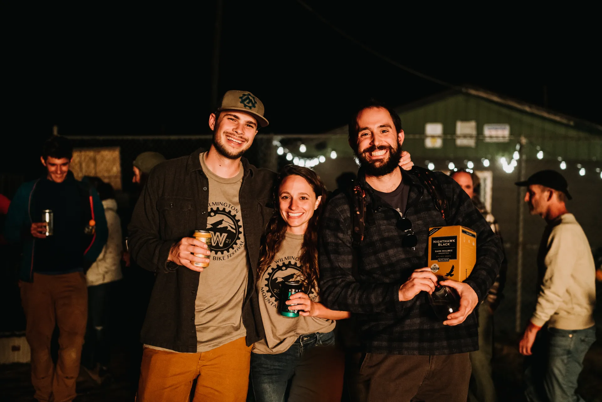 Three people at a nighttime outdoor event with BETA shirts holding beverages smiling