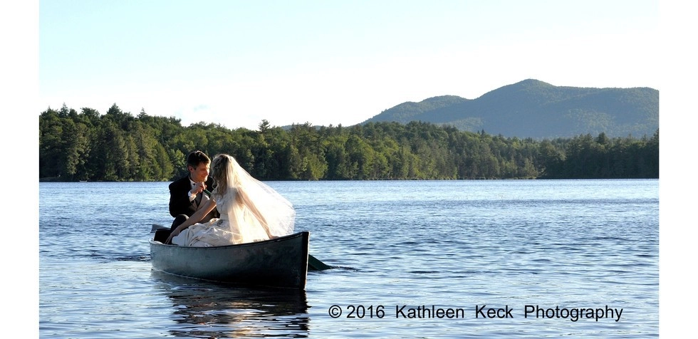 A bride and groom sit in a canoe on the water with mountains in the background.