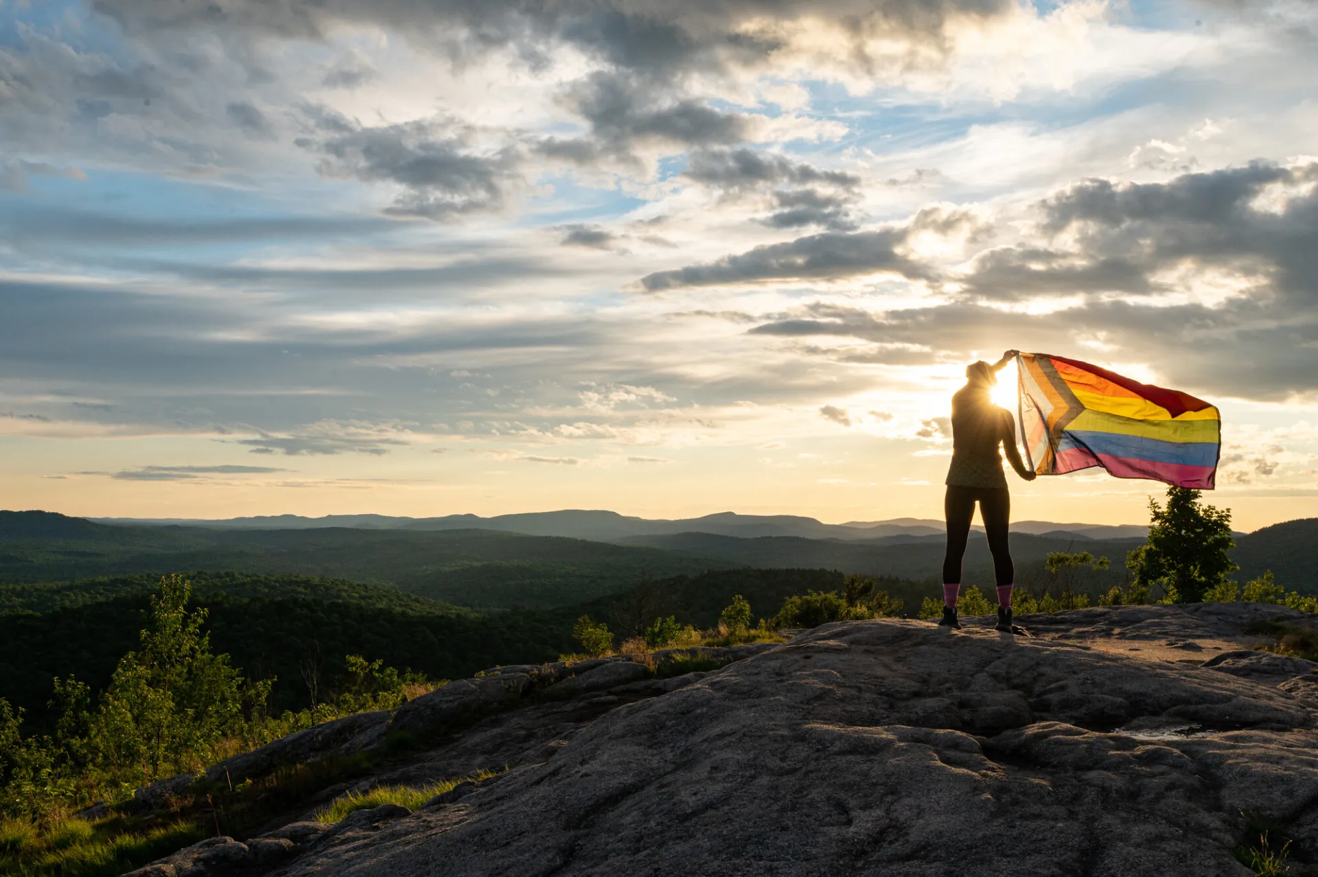 A person stands on a rocky hilltop, holding a brightly colored pride flag against a scenic sunset. The sky is dramatic with scattered clouds.