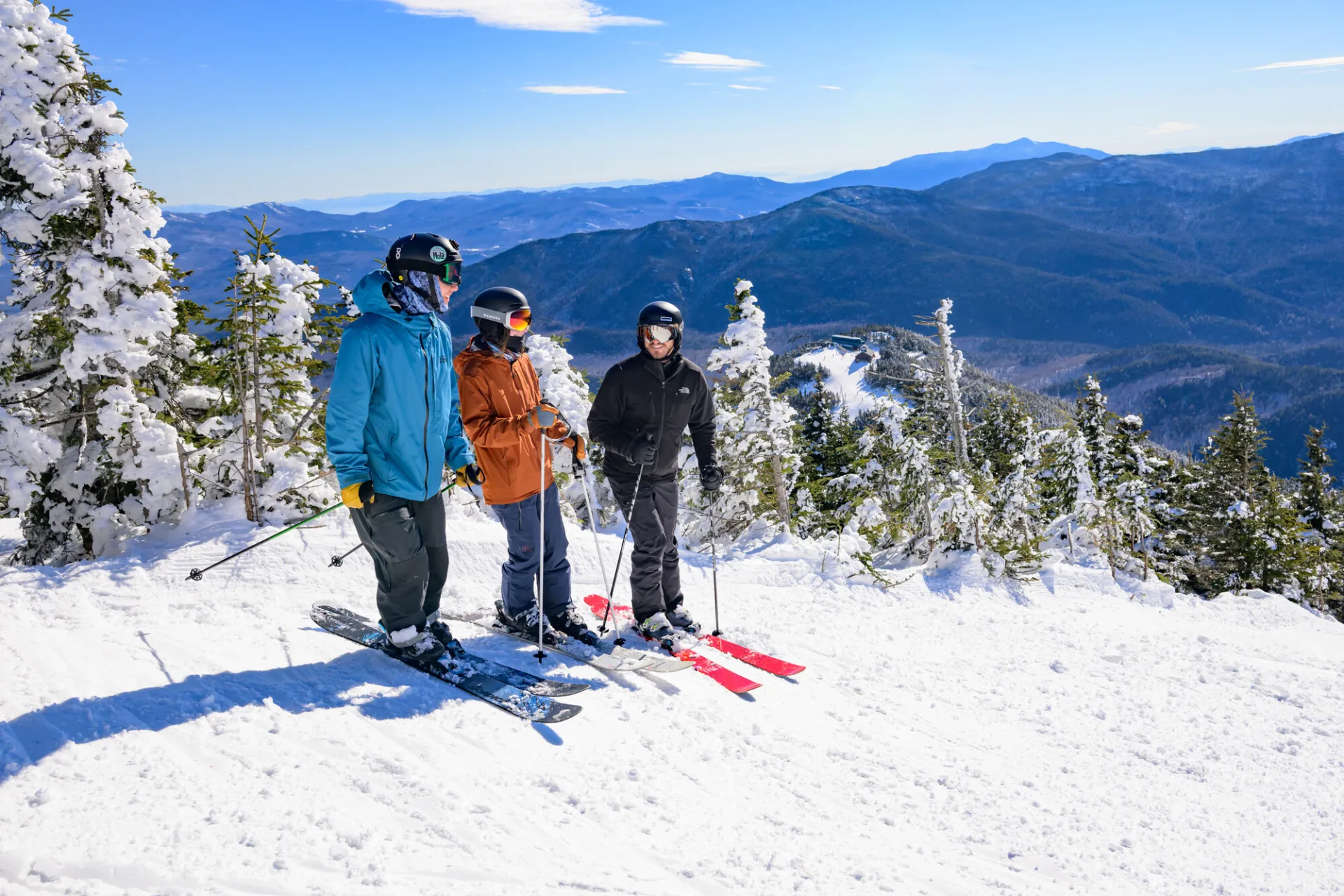 Three skiers stand at side of ski run in ski gear talking with snow encrusted trees beside them and snowy mountains in the distant background