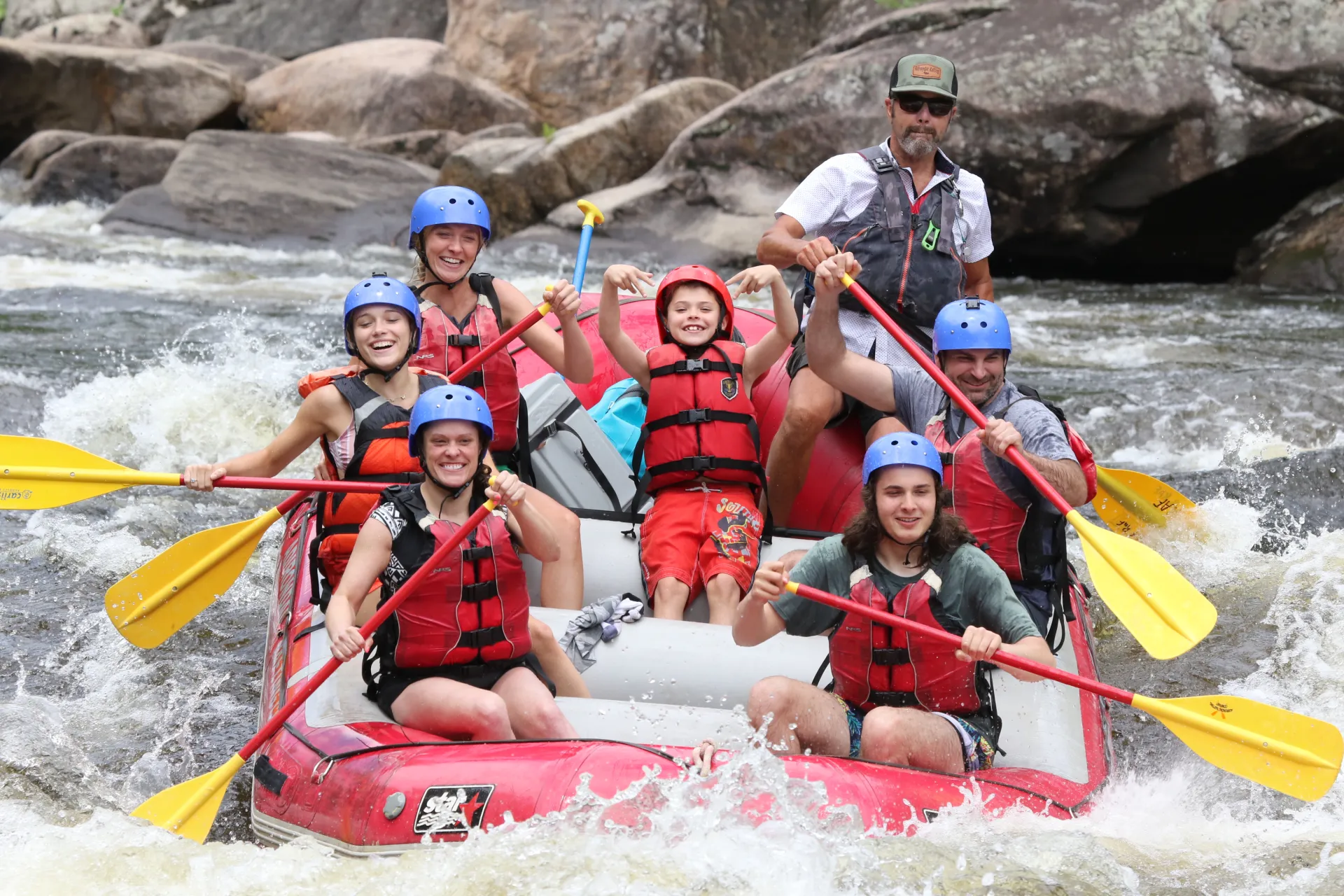 A smiling young man in a raft surrounded by friends waves at the camera.