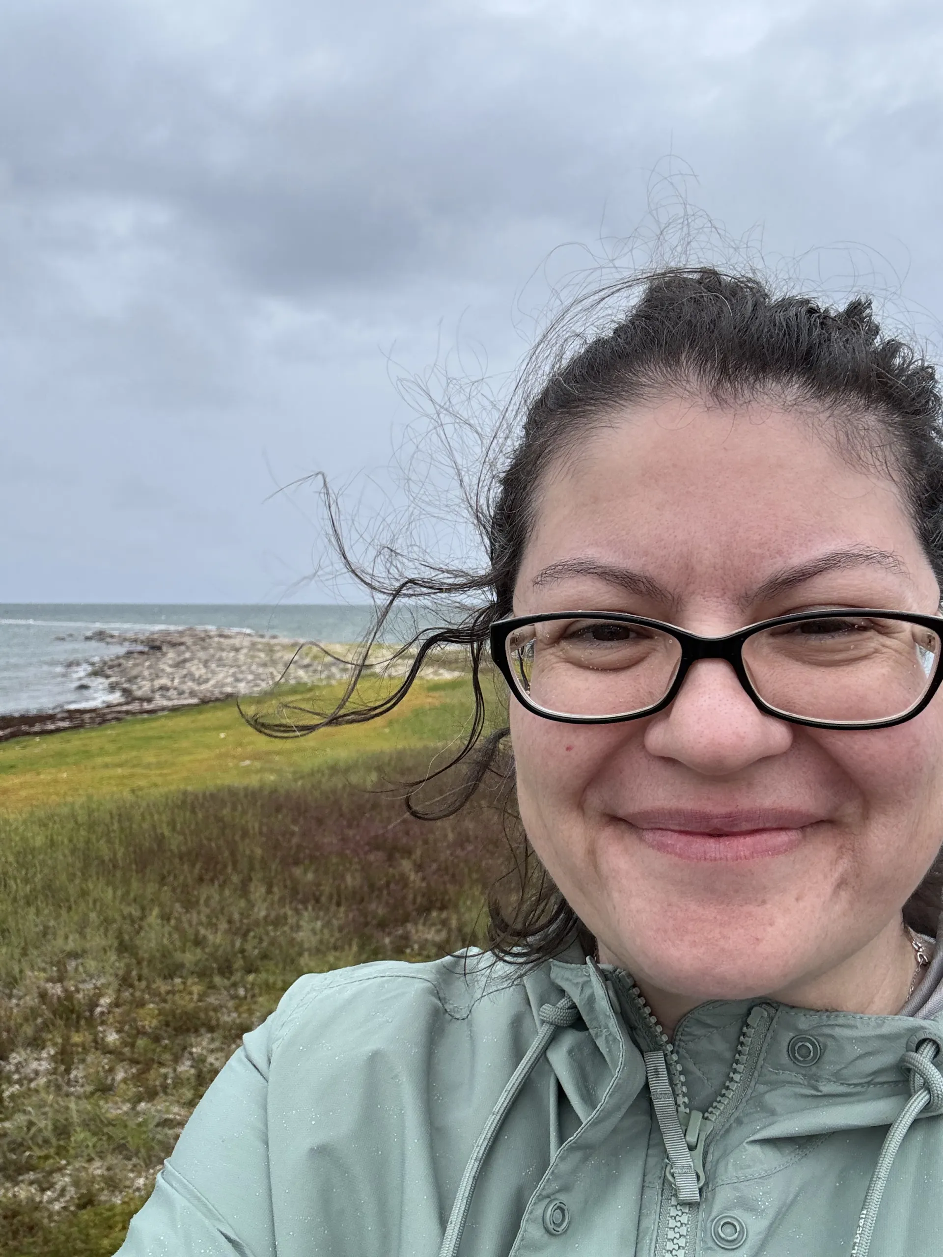 A woman with windblown hair smiles in front of a grey coastline.