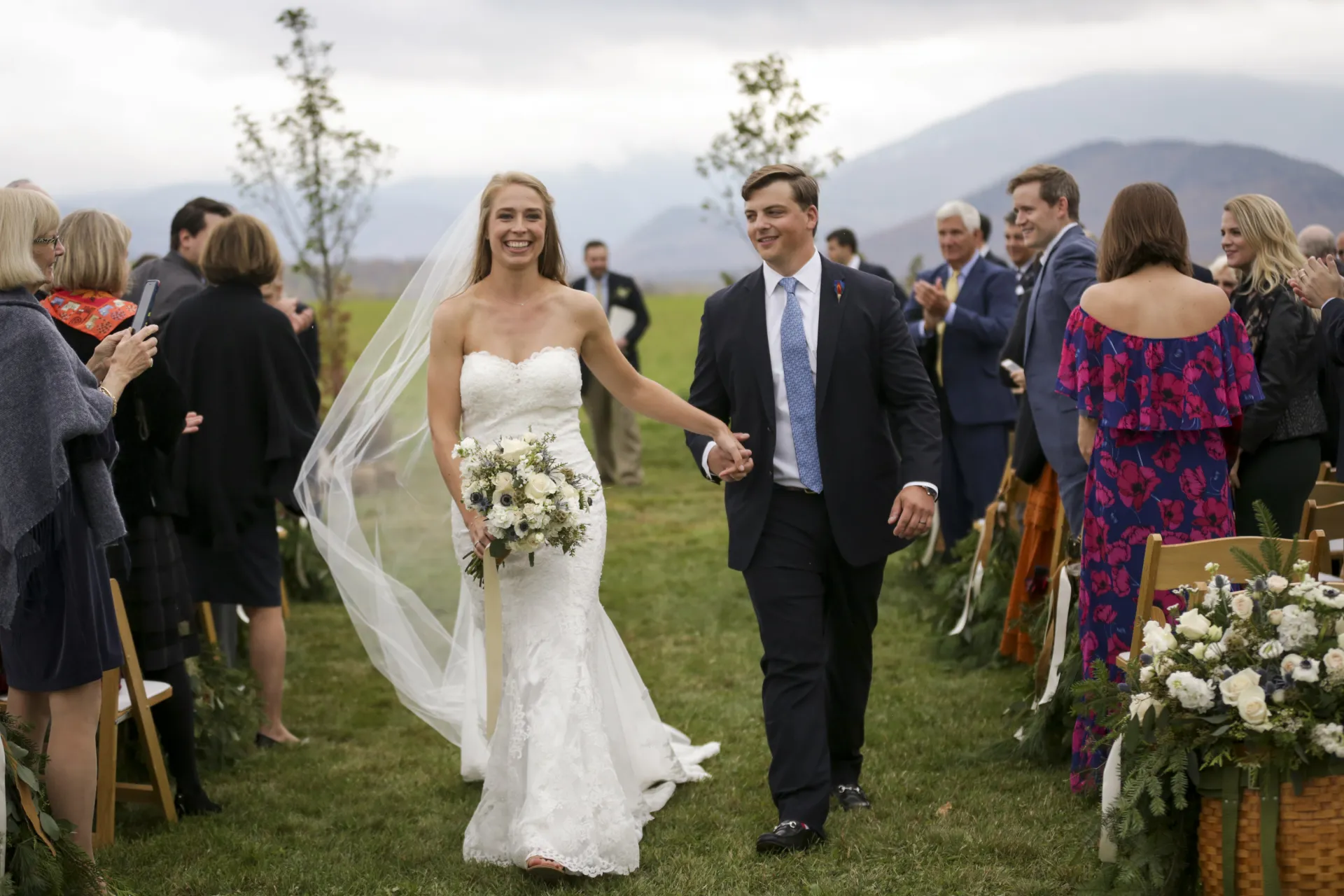 A bride and groom walk away from the alter while holding hands and smiling at friends and family and the mountain backdrop