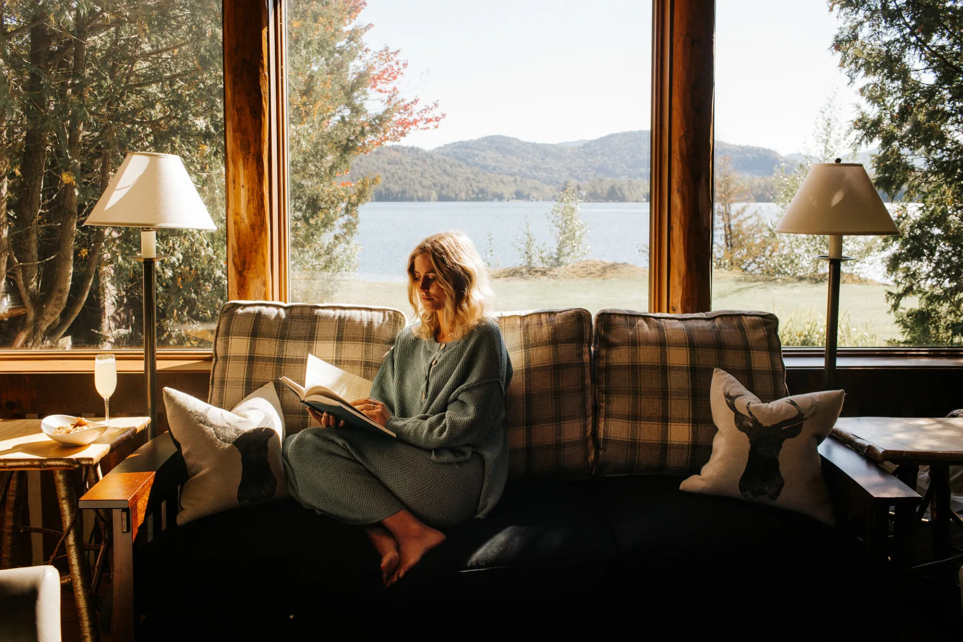 Blonde woman sits on couch with flannel print in loungewear reading a book in front of a window with lake and mountain view through window