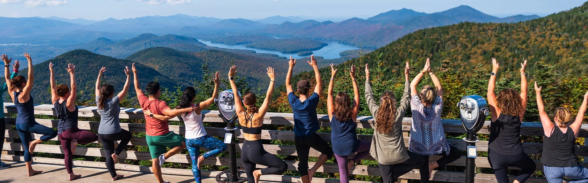 People holding tree pose on the deck at the top of Little Whiteface Mountain overlooking the surrounding mountains and Lake Placid.