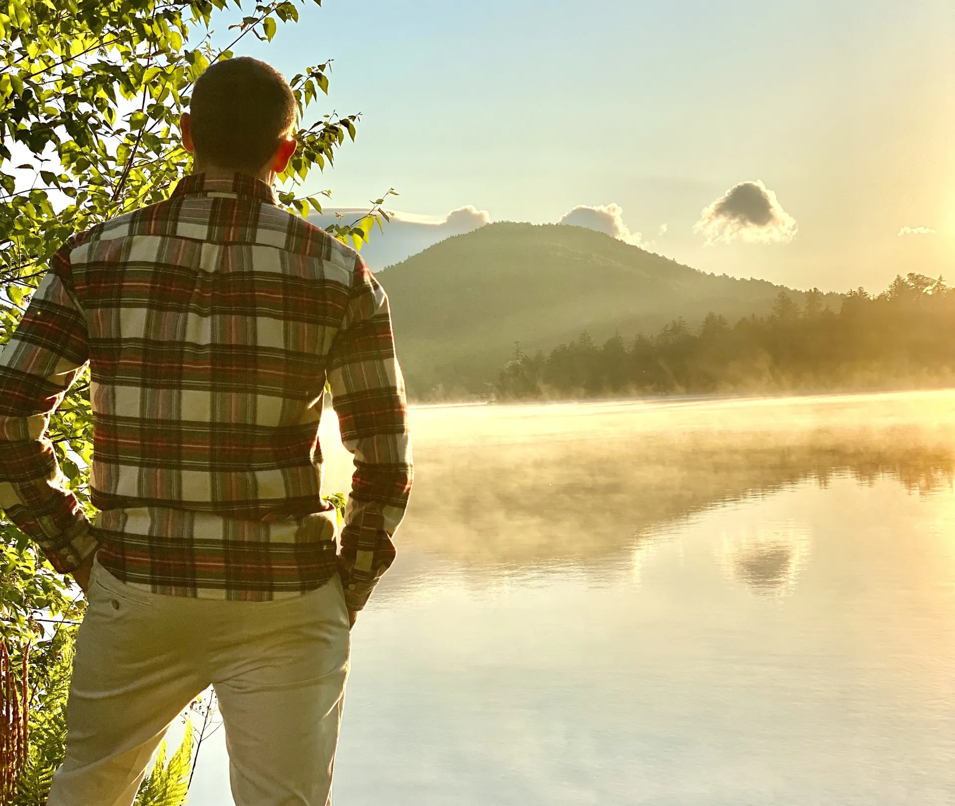 A man in a flannel looks out over a misty lake and mountains.