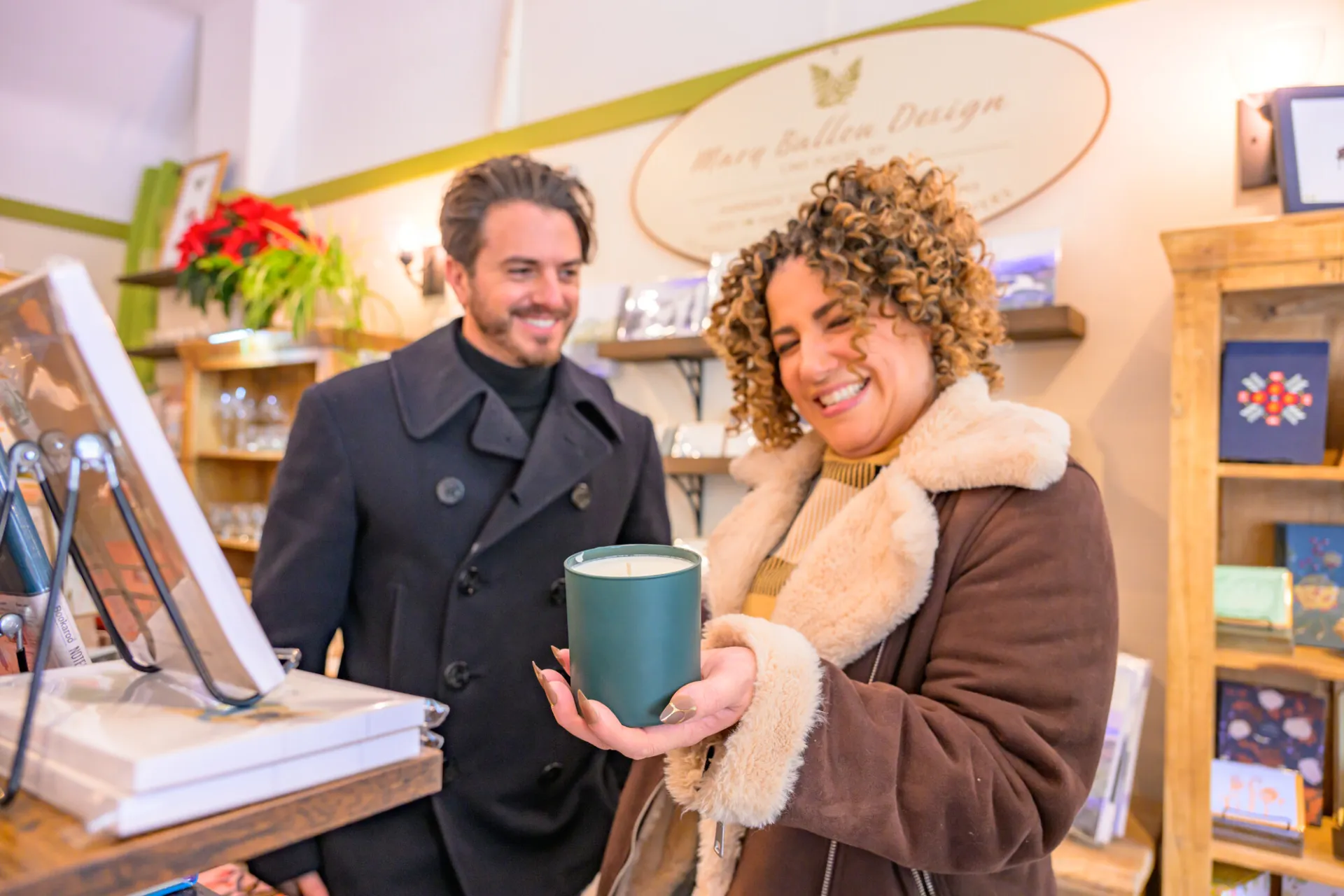 A man and a woman in winter coats smile as the woman holds a candle in a small, quaint gift shop.