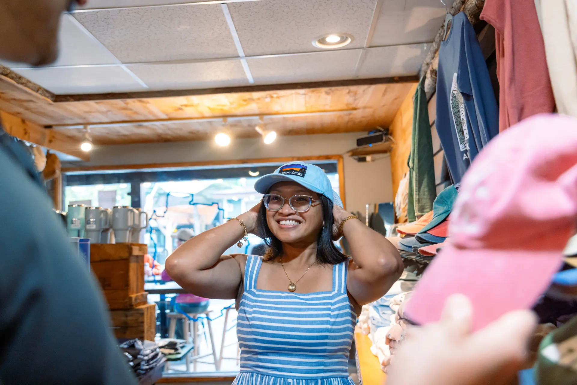 A smiling woman wearing a blue-and-white striped dress and glasses tries on a light blue baseball cap inside a shop.