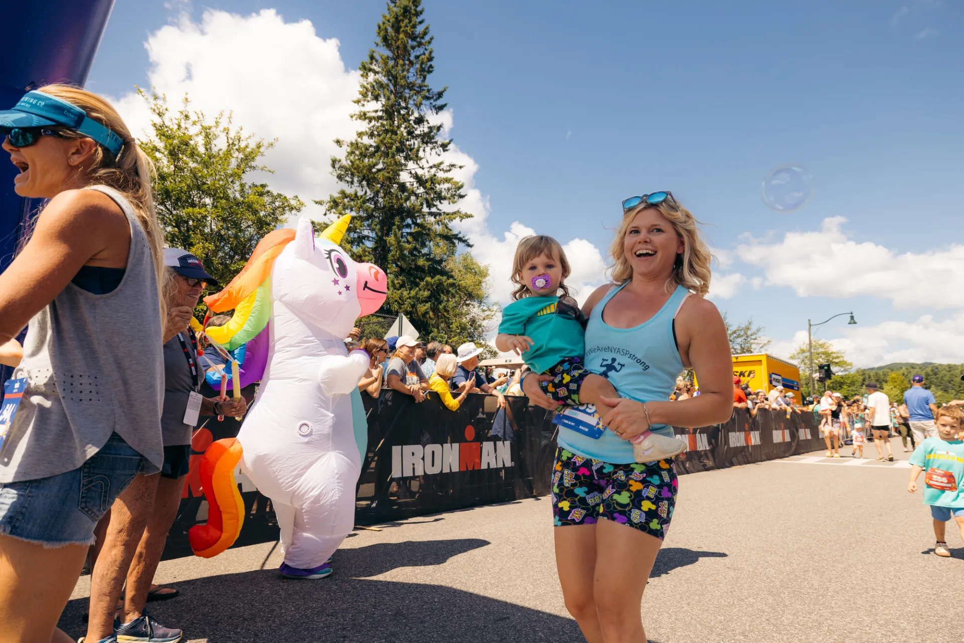 Woman in tank top and colorful shorts holds toddler while walking across finish line with inflatable unicorn behind