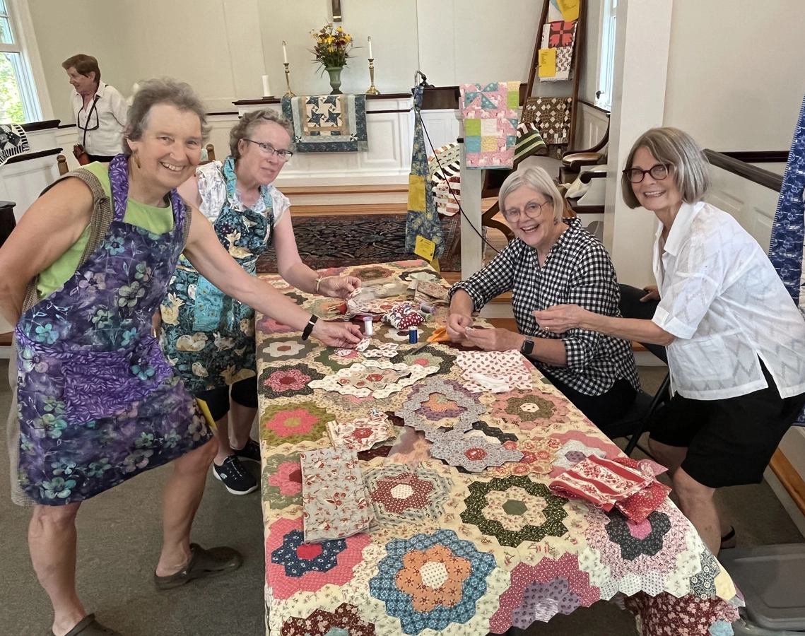 Four senior woman gathered around a table covered in a quilt and quilting materials look at camera smiling