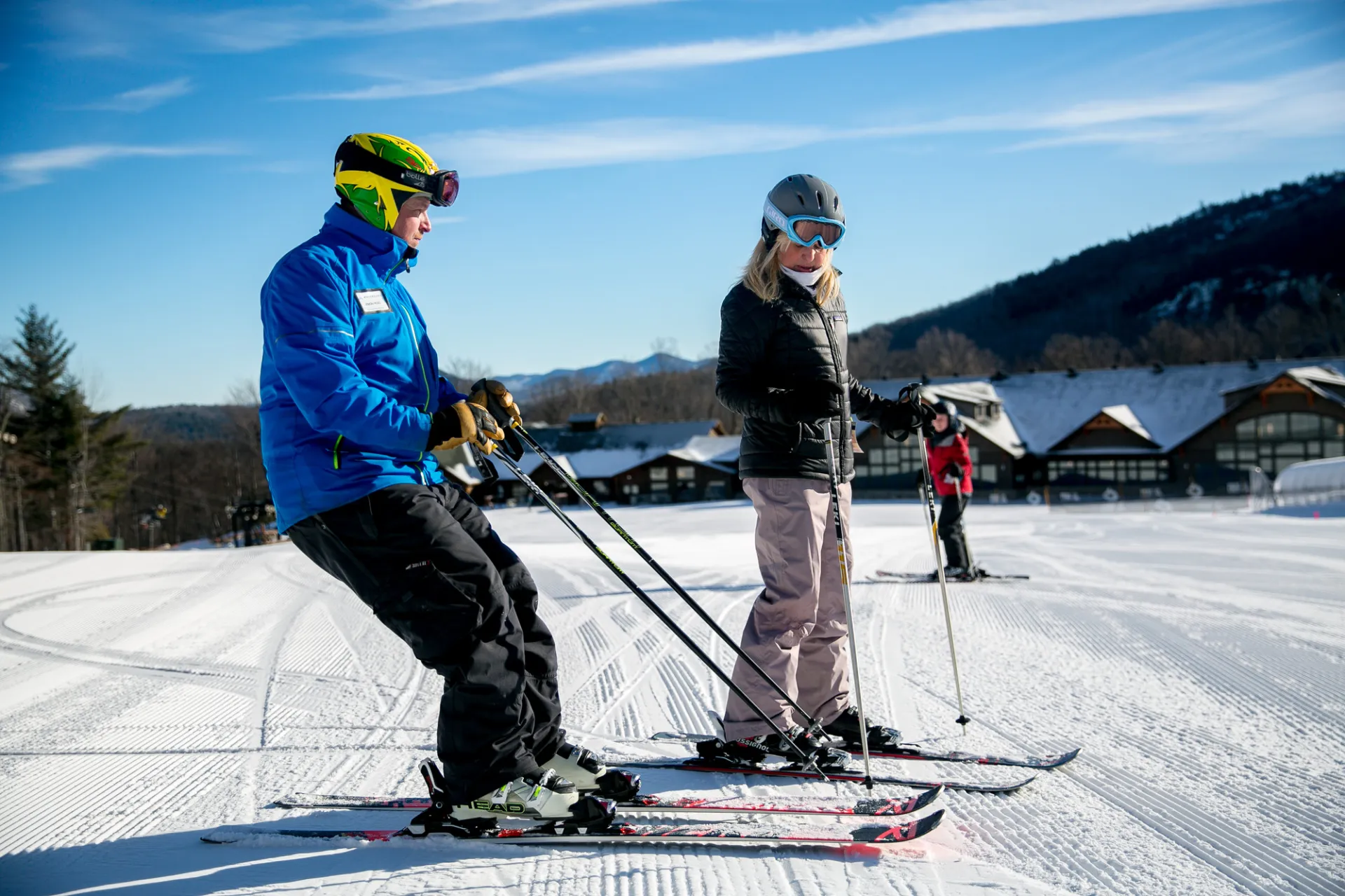Ski instructor on skis in blue coat shows new female skier on skis how to lean back in your boots