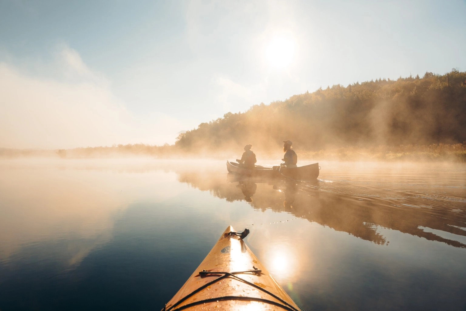 Two people paddle canoe on mirror-like water with fog seen past bow of orange kayak
