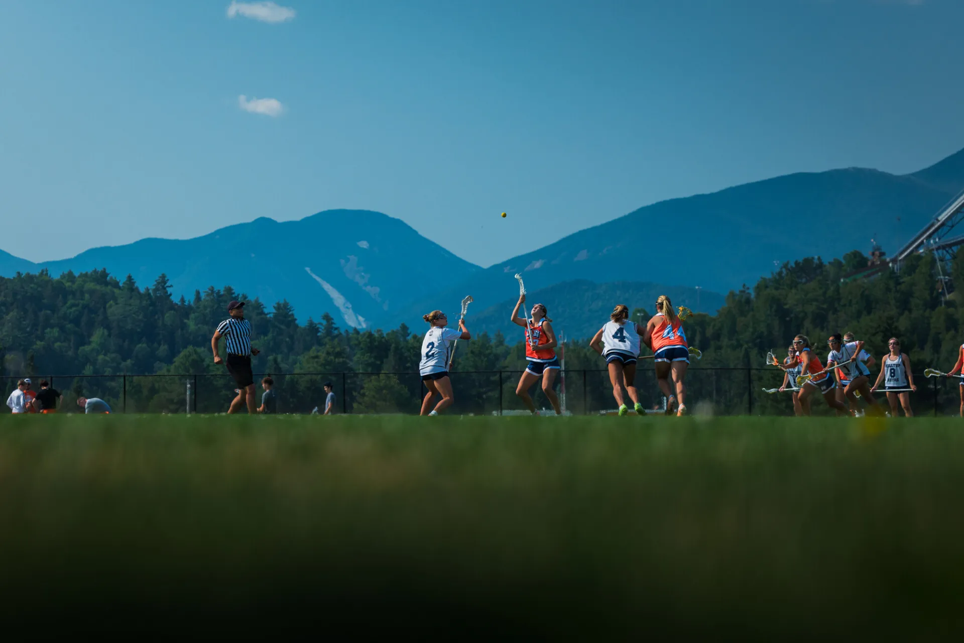 Two women holding lacrosse sticks look up at yellow lacrosse ball in the air during play on field with mountain views