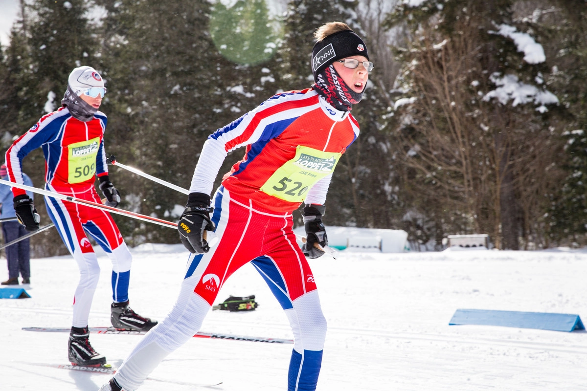 A nordic skier in red, white, and blue cross-country ski gear and a yellow race bib skis past the camera from left to right on a side snowy trail in the woods.
