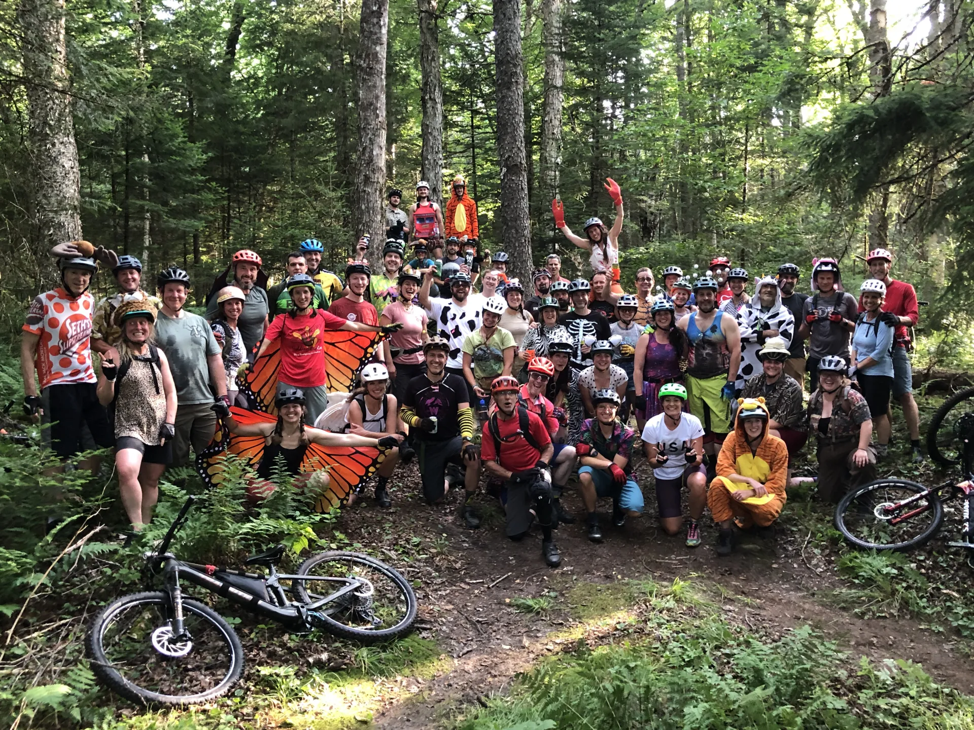 Large group of people, many dressed in a combo of animal costumes and biking gear, pose for photo in the woods with bikes